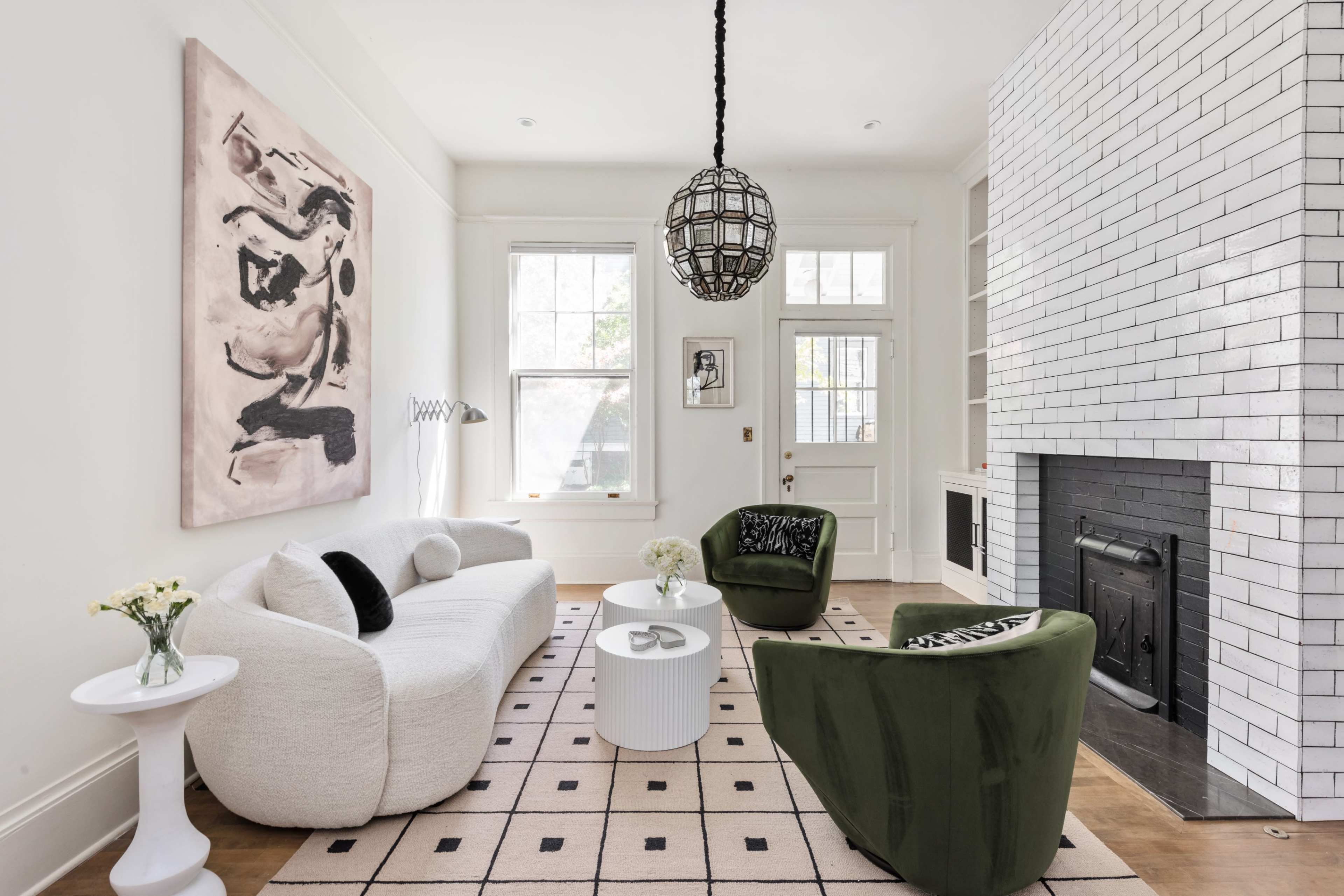 The room features a white sofa, two green chairs, a round coffee table, and a black-and-white tiled fireplace, with large windows and an abstract painting on the wall.