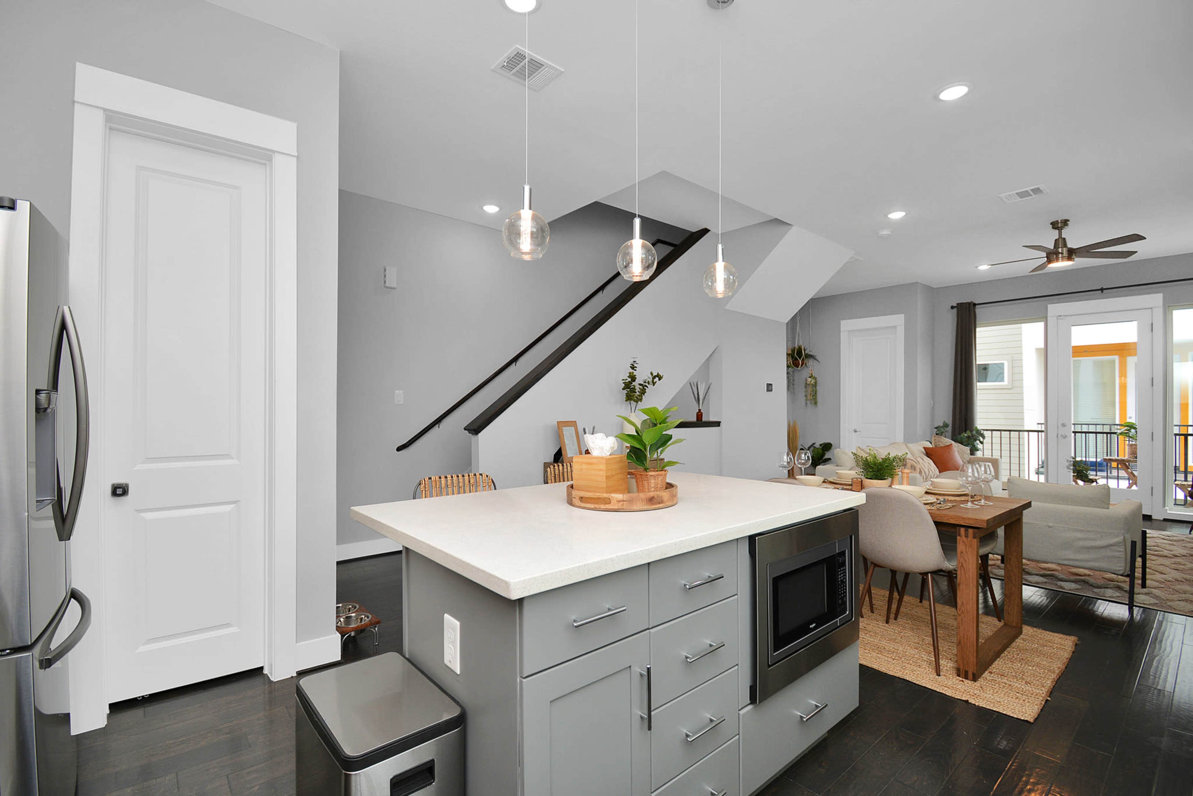 A modern kitchen features a central island with seating, open shelving, and leads to a dining area with a staircase in the background.