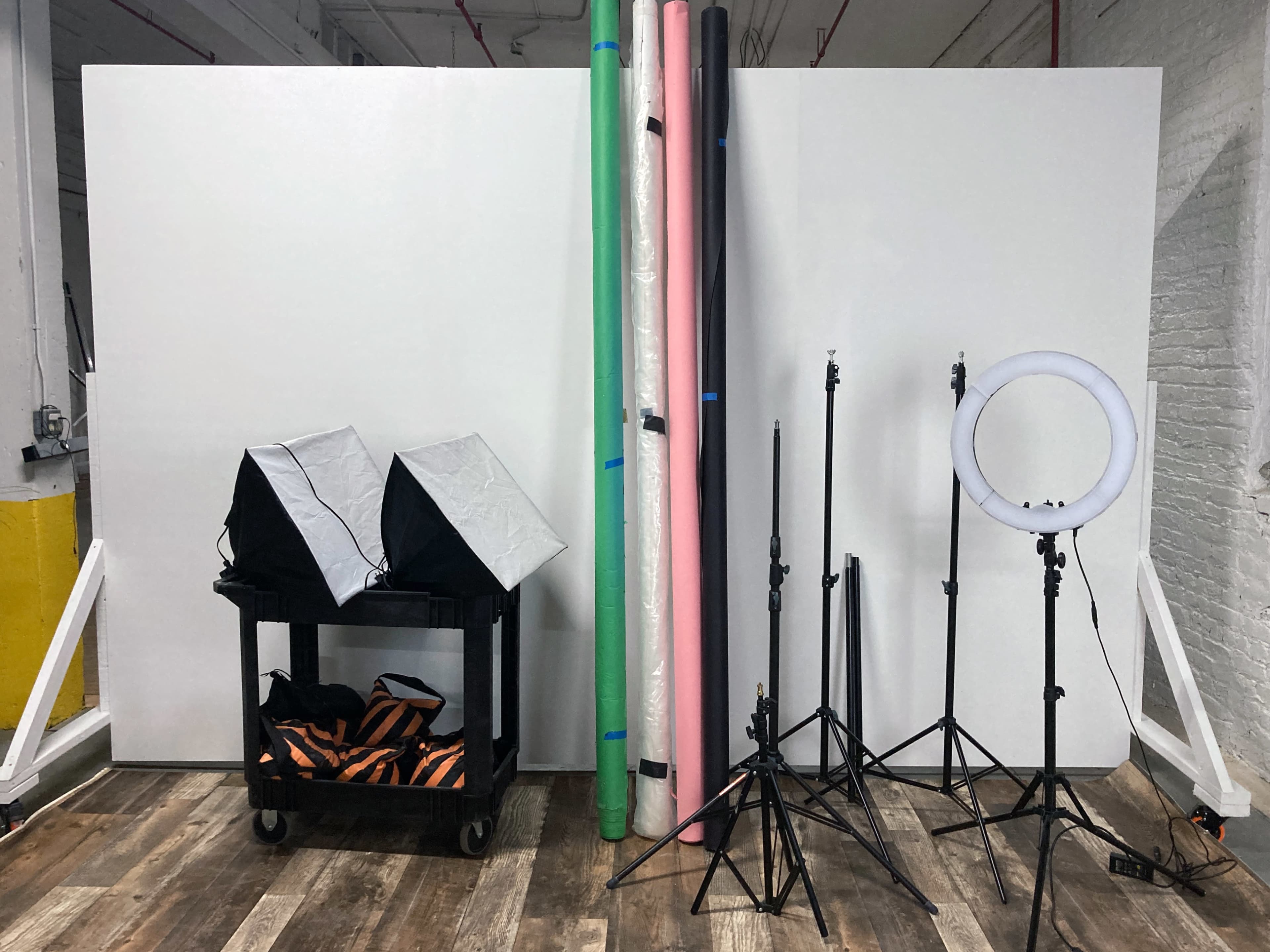 A photography setup with two softbox lights, a cart with equipment, and multiple backdrop rolls leaning against a wall.