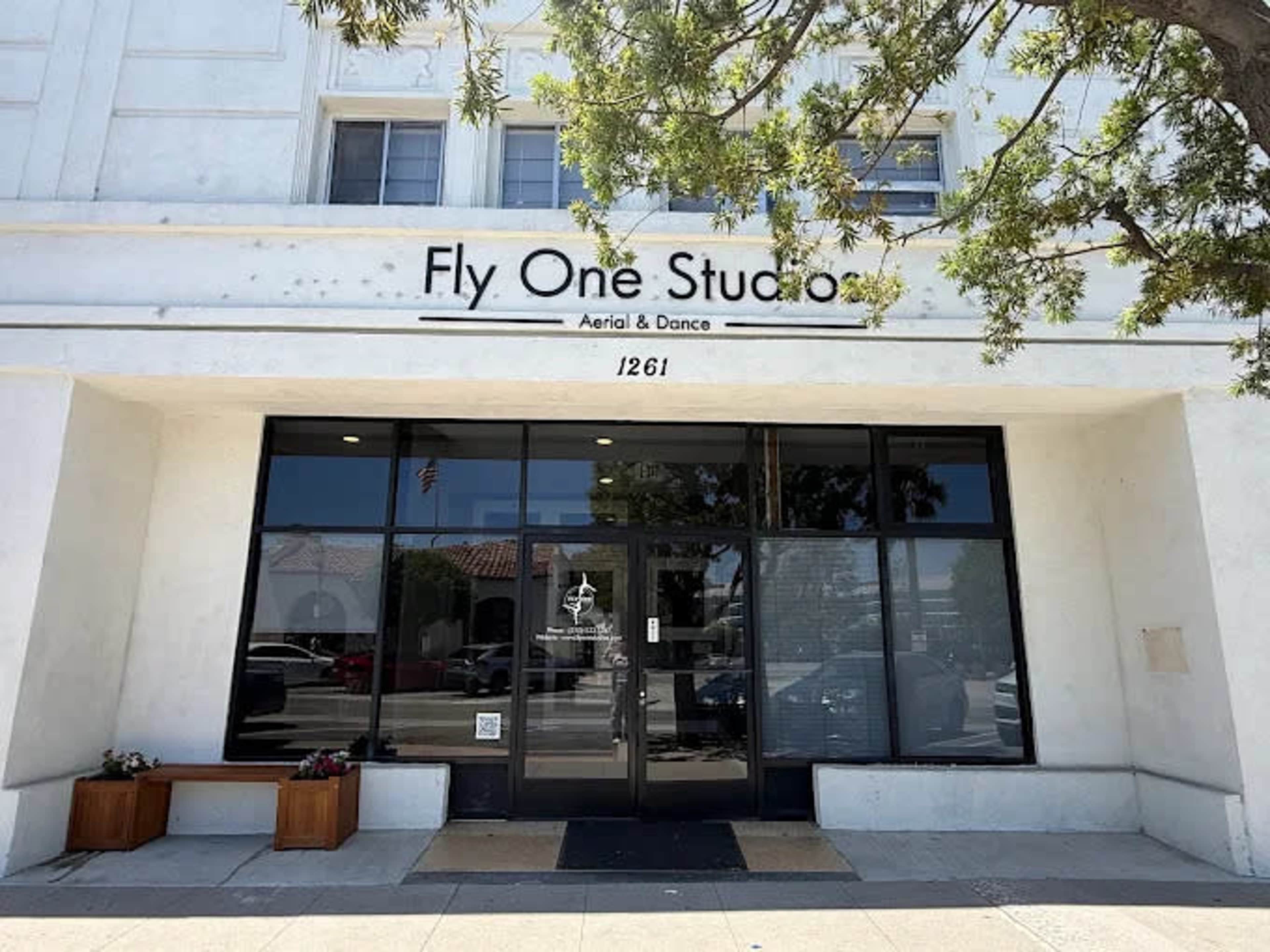 The image shows a building facade for "Fly One Studios" with large glass doors and a sign indicating it offers aerial and dance classes.