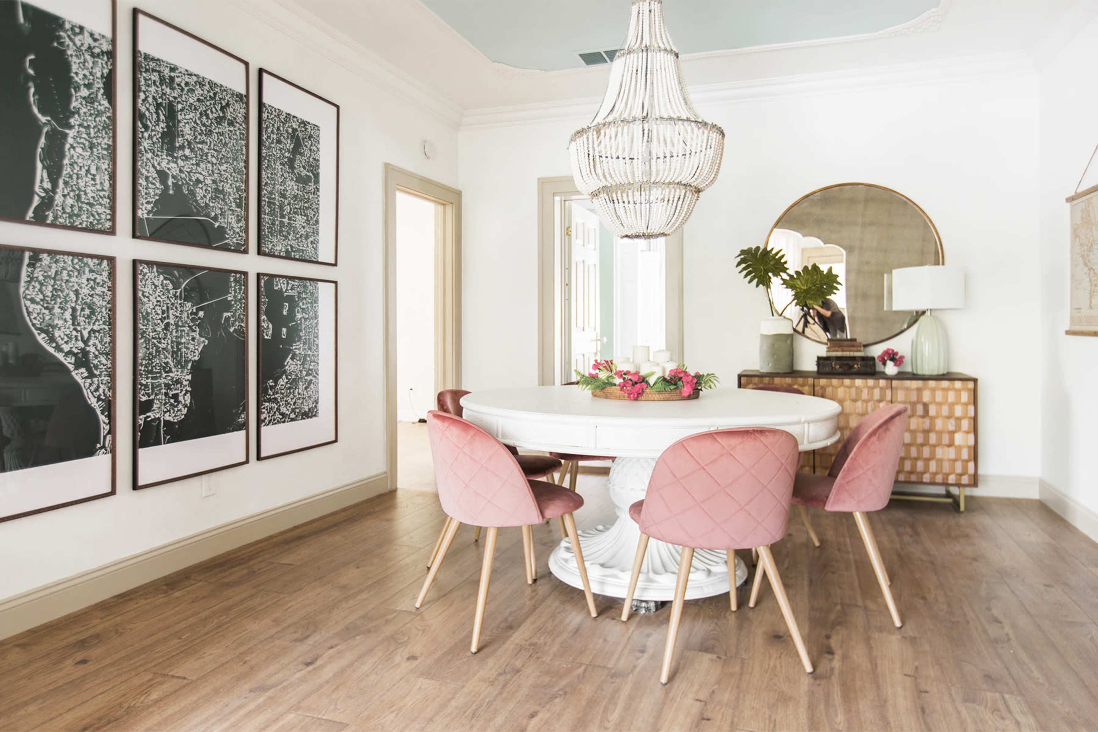 A circular dining table surrounded by pink chairs is set in a modern room featuring large framed maps, a large chandelier, and a mirror.