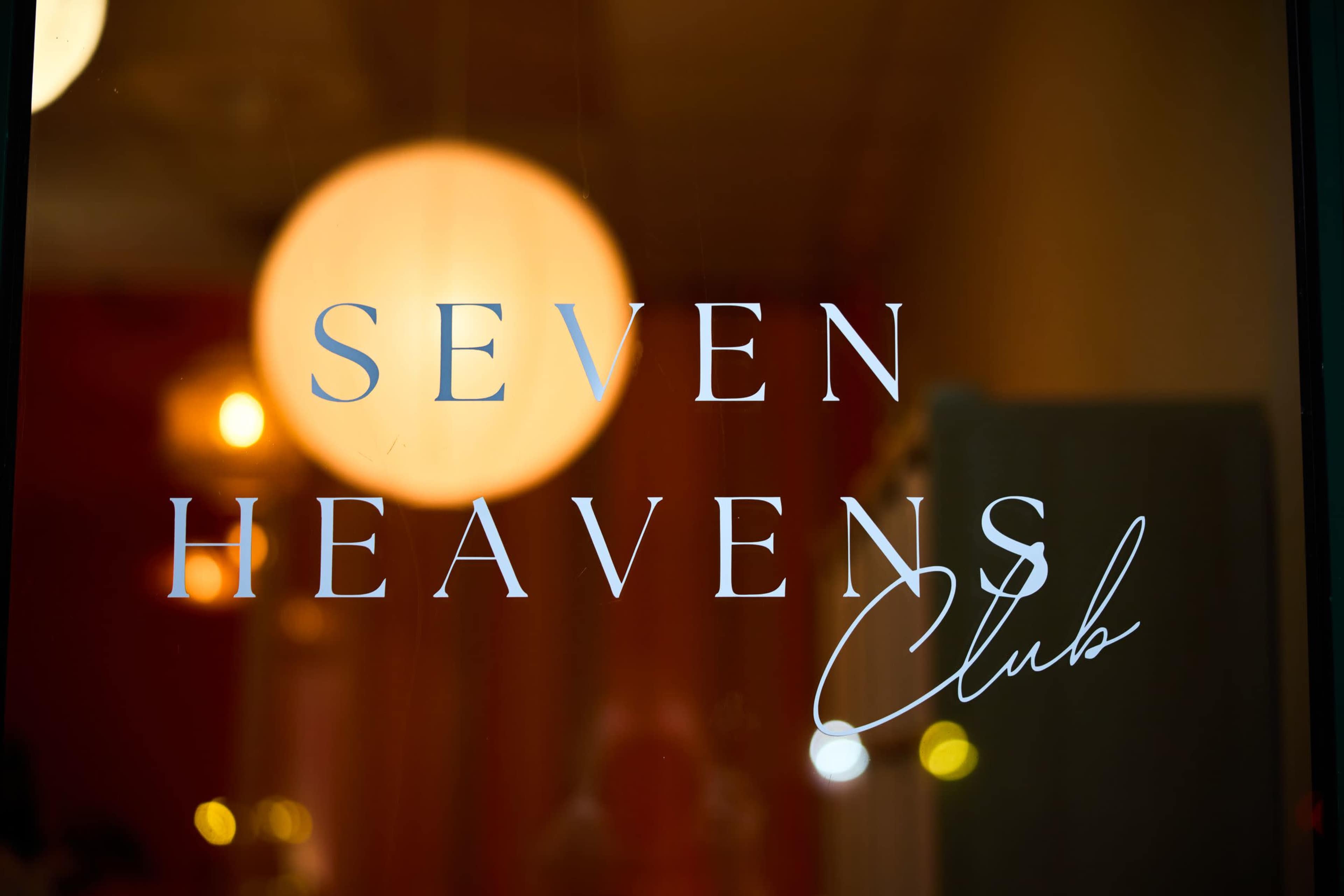The image shows the words "SEVEN HEAVENS Club" displayed on a glass surface, with soft, glowing lights in the background.