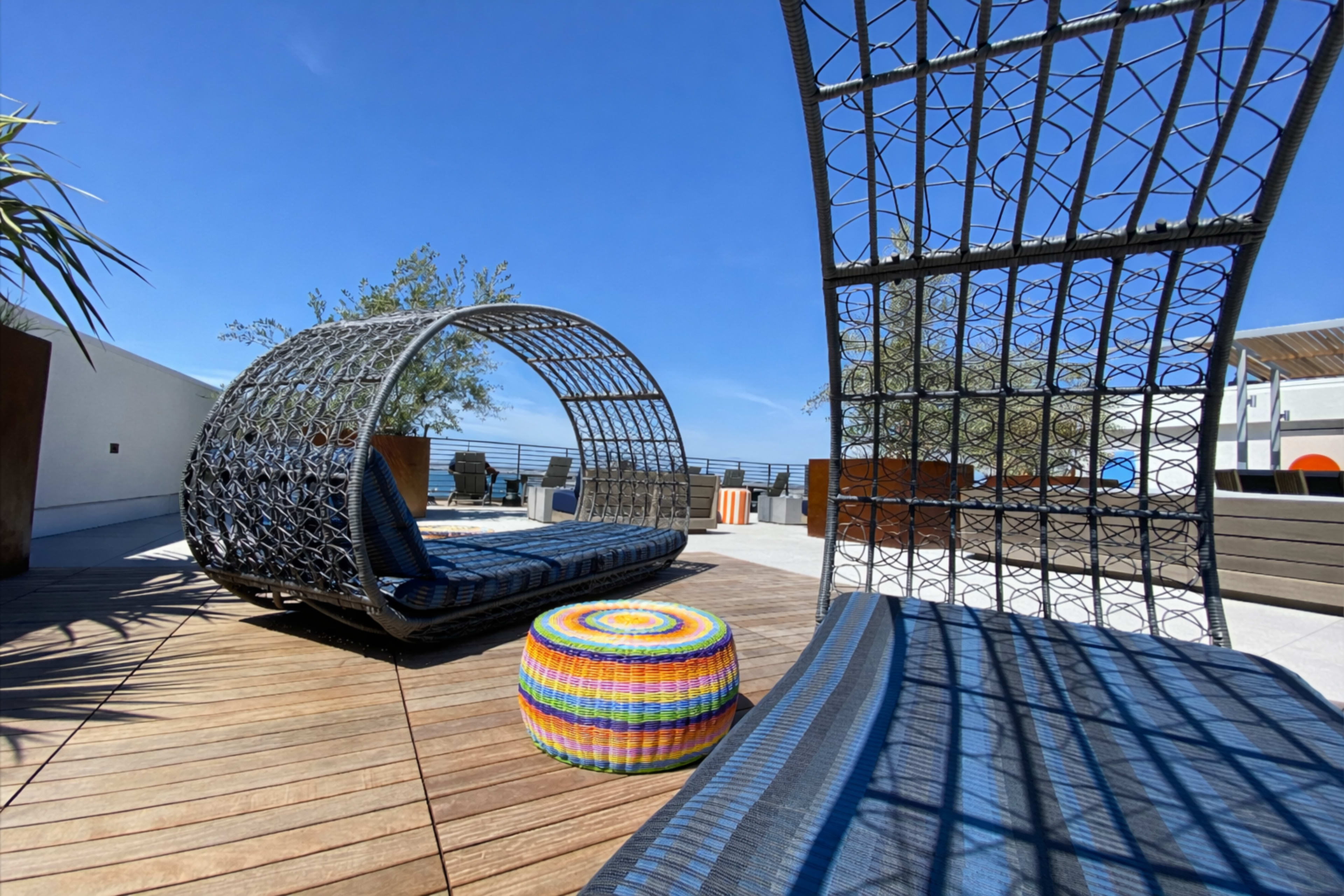 The image shows a rooftop lounge area featuring a circular wicker chair, a colorful pouf, and a wooden deck under a clear blue sky.