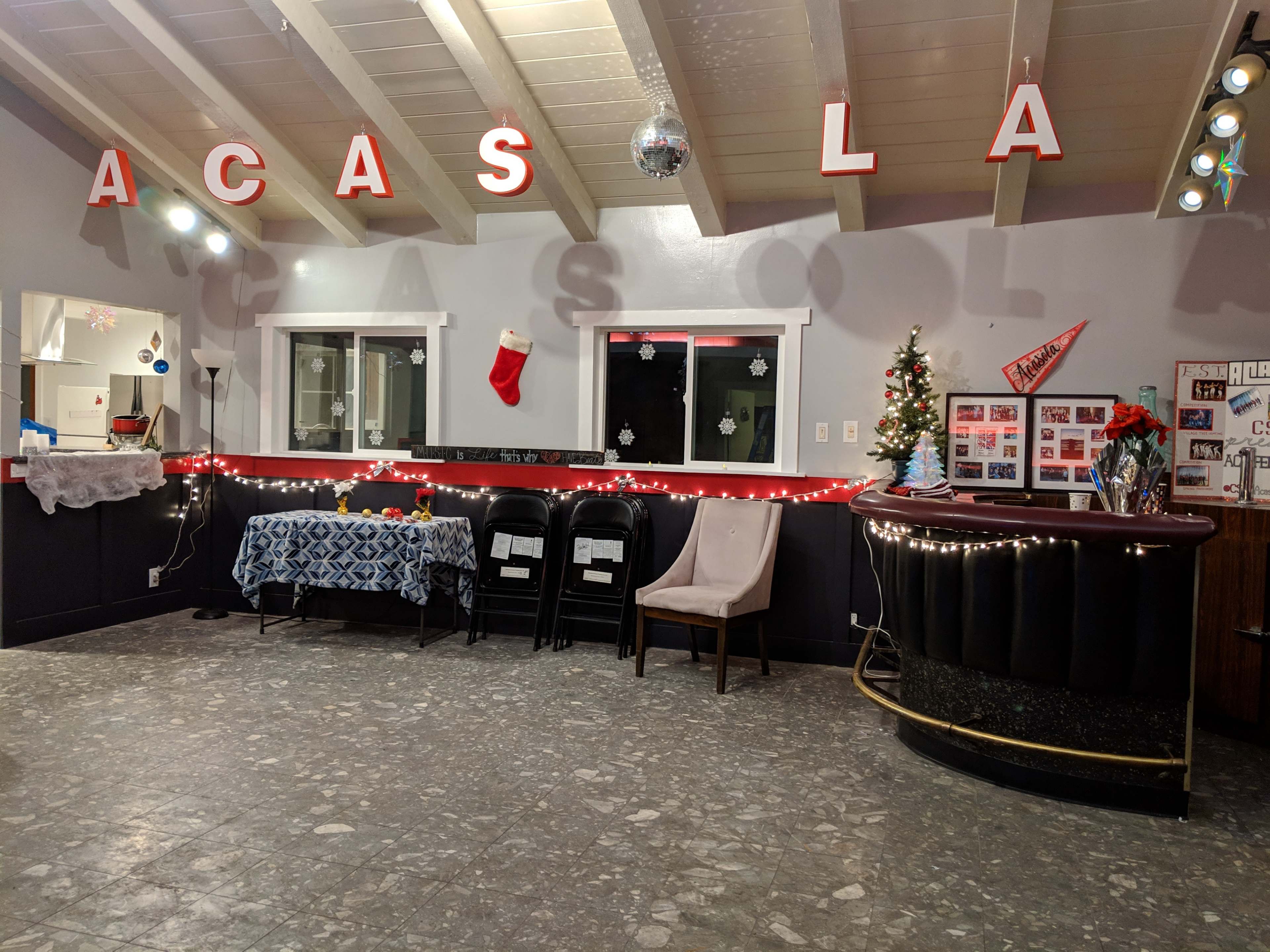 The interior of a warmly lit room features a decorated bar, a table covered with a blue cloth, black chairs, and festive decorations, including a Christmas stocking.
