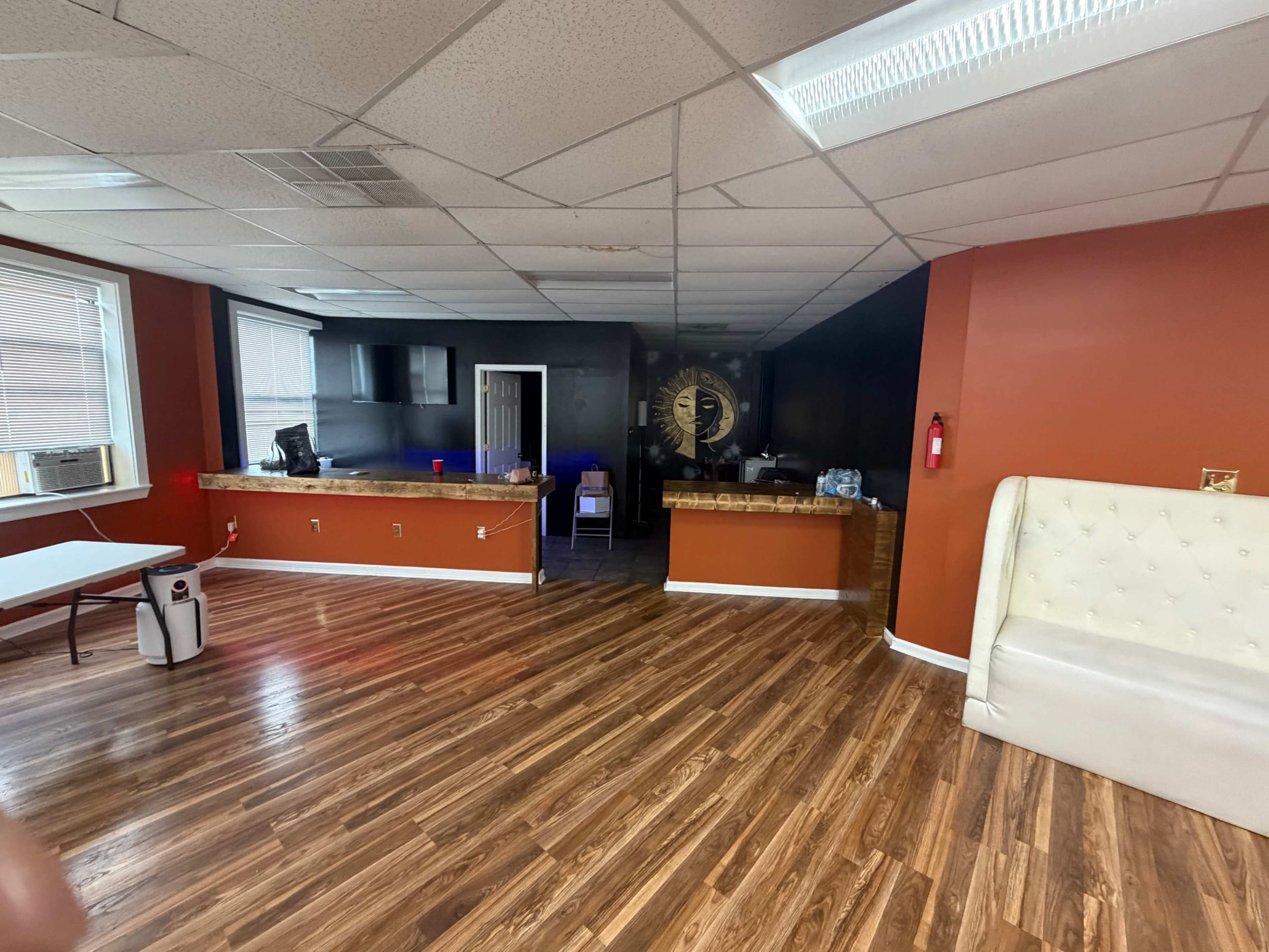 The image shows a spacious indoor area with a wooden floor, featuring a reception desk in the background and a white sofa against the wall.