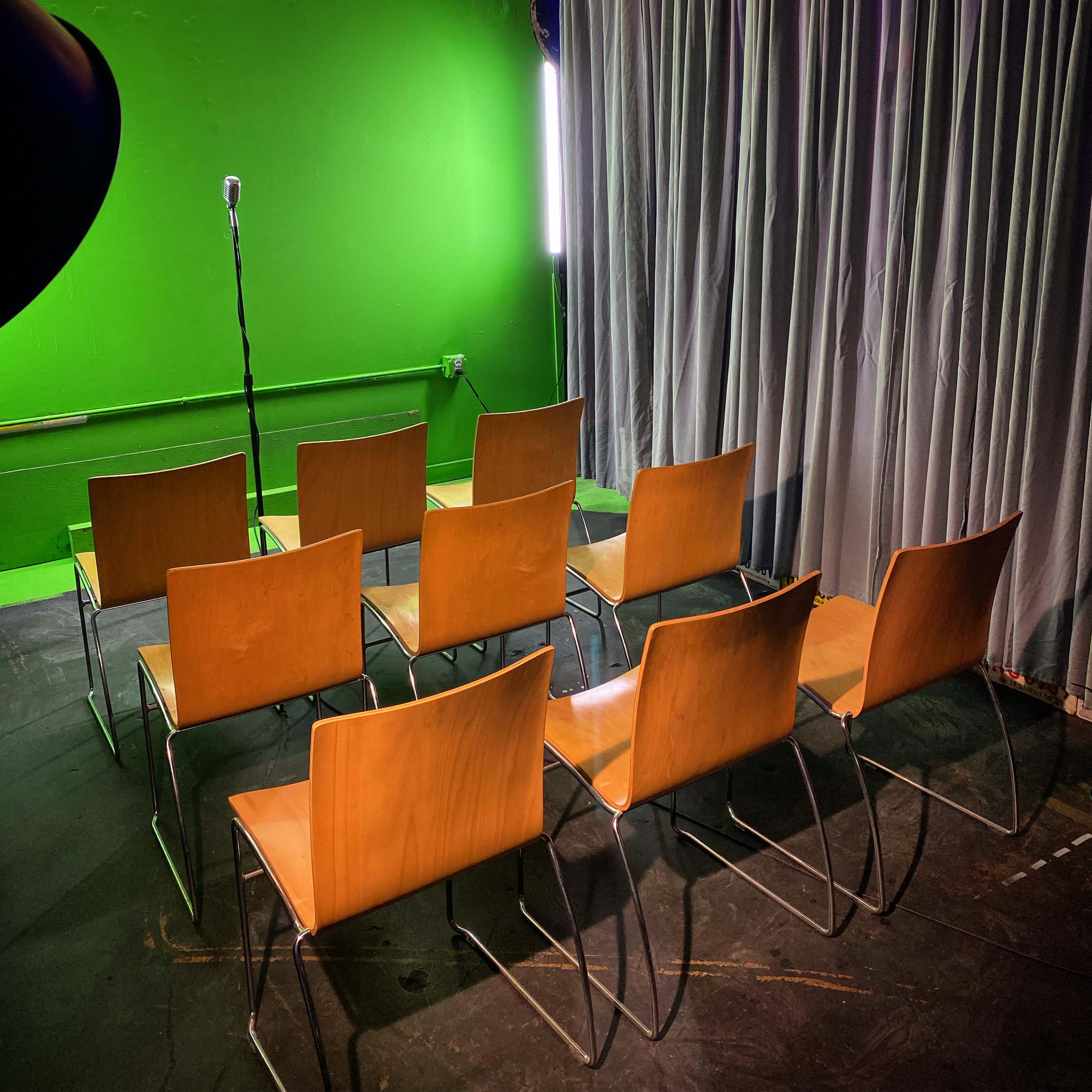 A row of wooden chairs positioned in front of a microphone, with a green wall and curtains in the background.