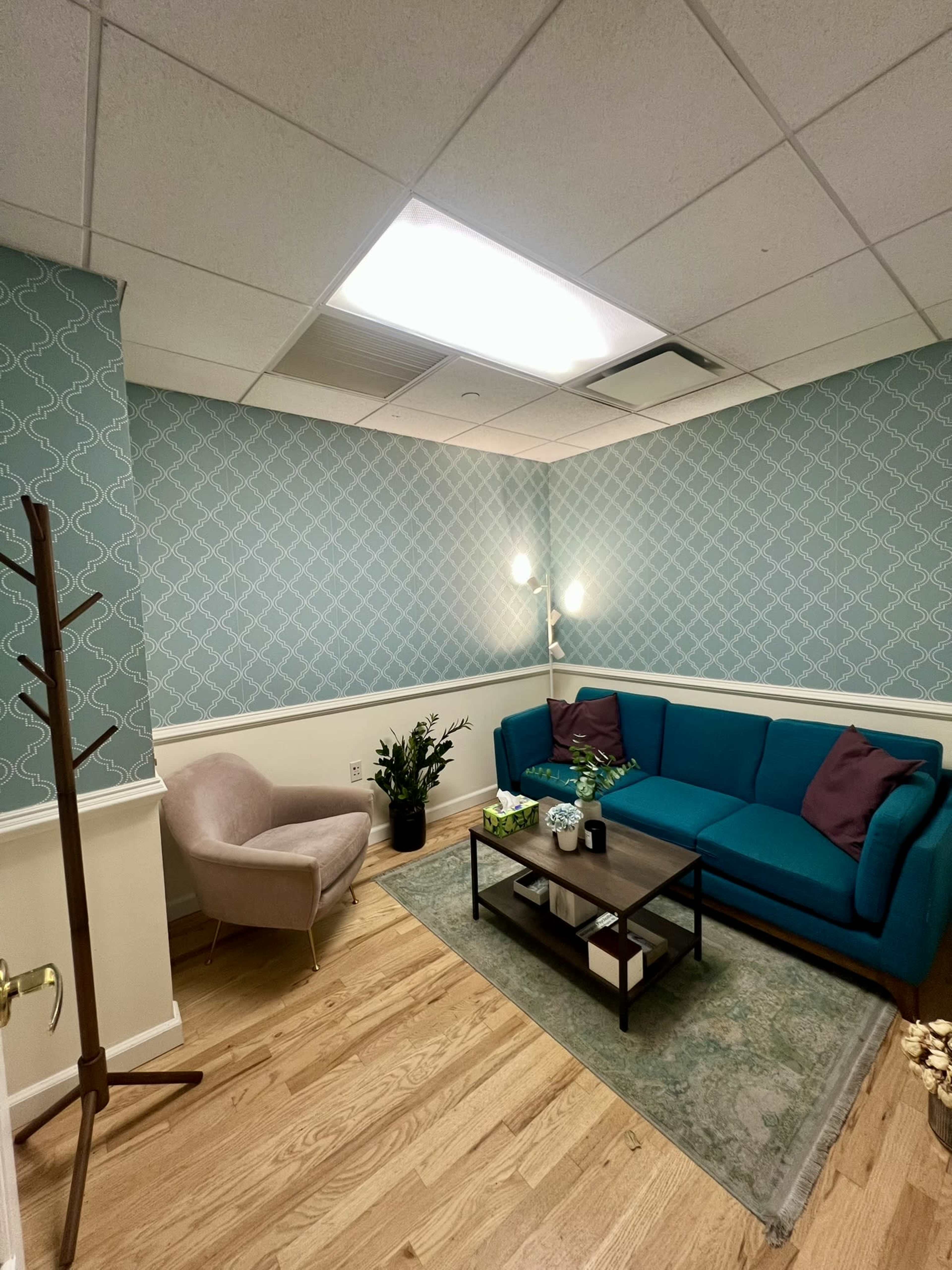 The image shows a small waiting area featuring a teal sofa with purple cushions, a light-colored armchair, a coffee table, and a decorative rug against patterned wallpaper.