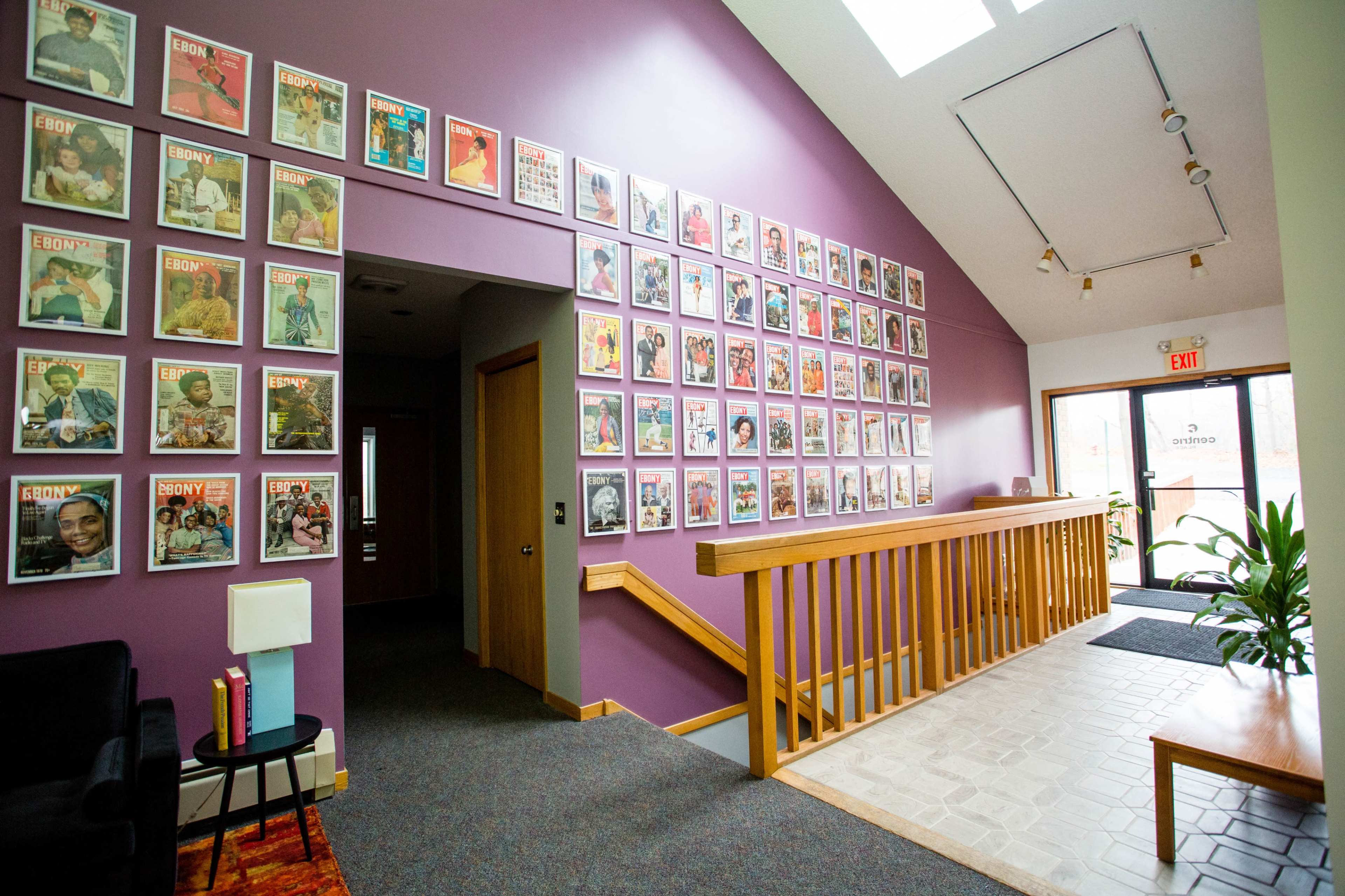 An interior hallway featuring a wall adorned with framed covers of Ebony magazine, alongside a wooden railing and a small seating area.