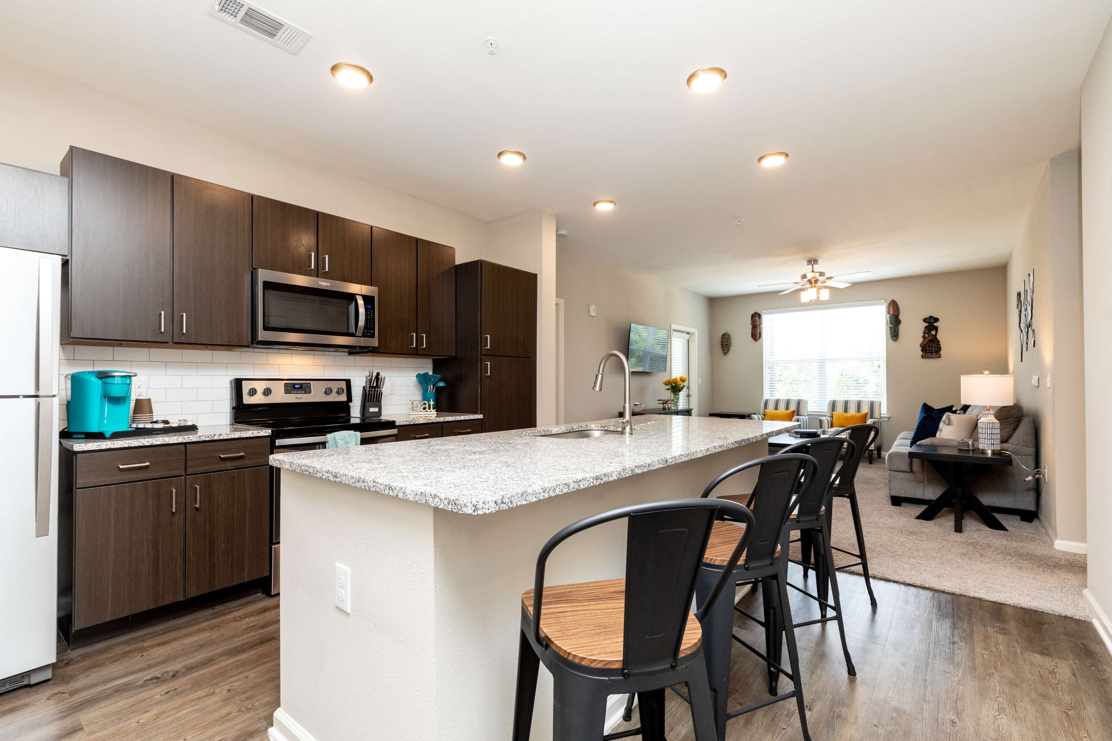 A modern kitchen with dark cabinets, a stainless steel refrigerator and microwave, and a center island with bar stools, leading into a living area with carpeted flooring.