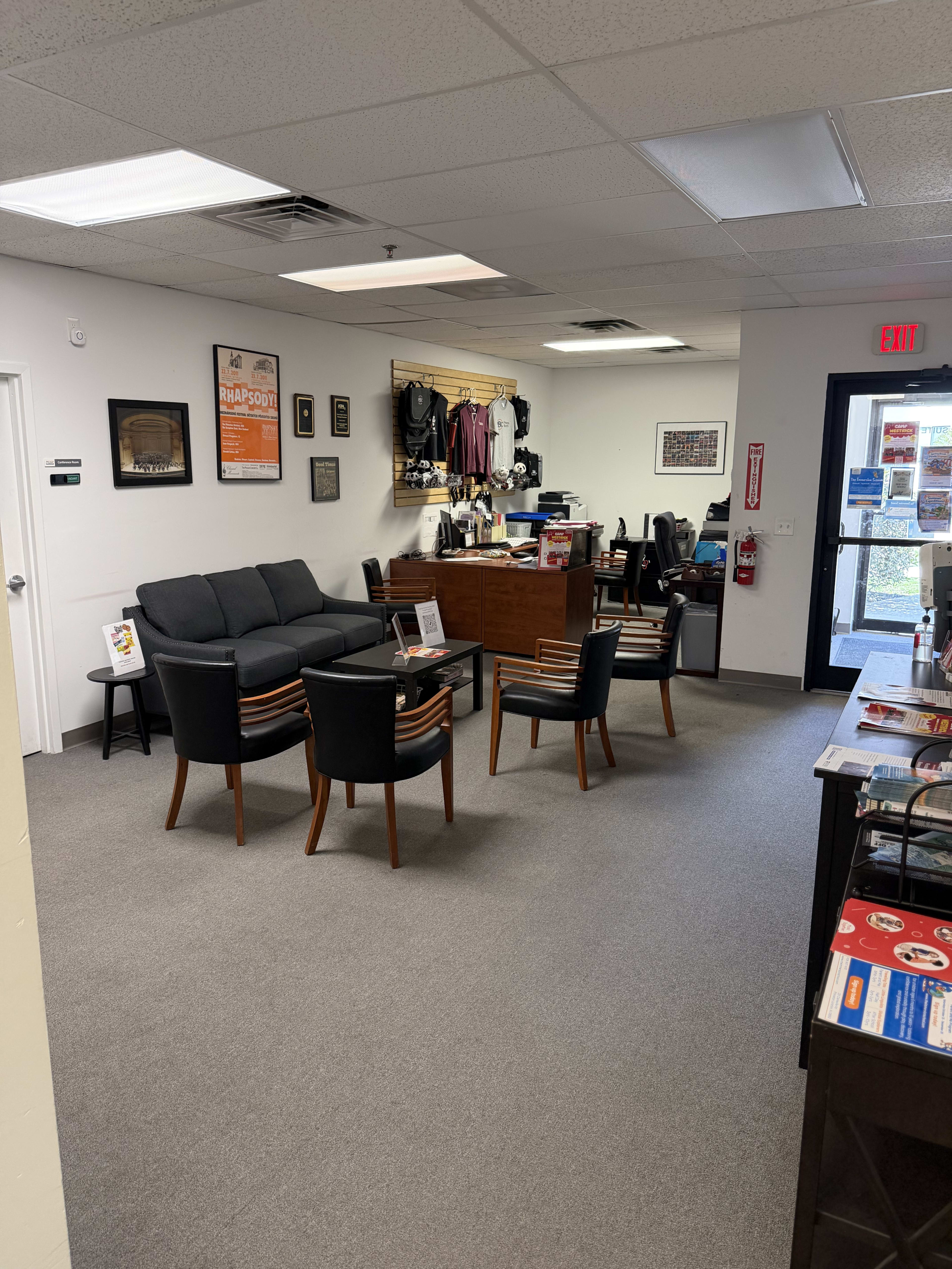 The image shows a waiting area with a black sofa and chairs arranged around a coffee table, a reception desk in the background, and various items displayed on the walls.