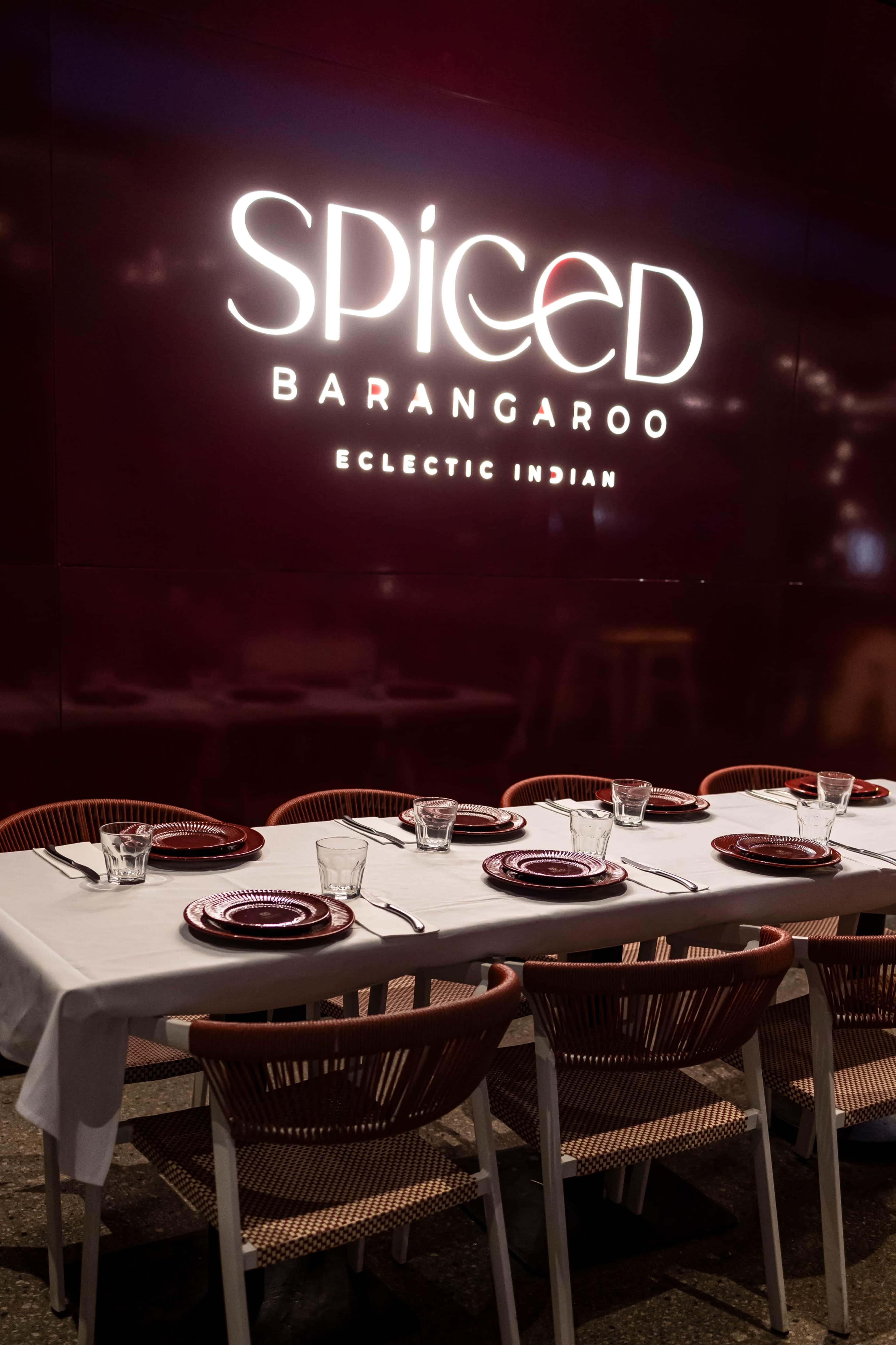 A dining table is set with red plates and glassware in front of a glowing restaurant sign that reads "Spiced Barangaroo Eclectic Indian."