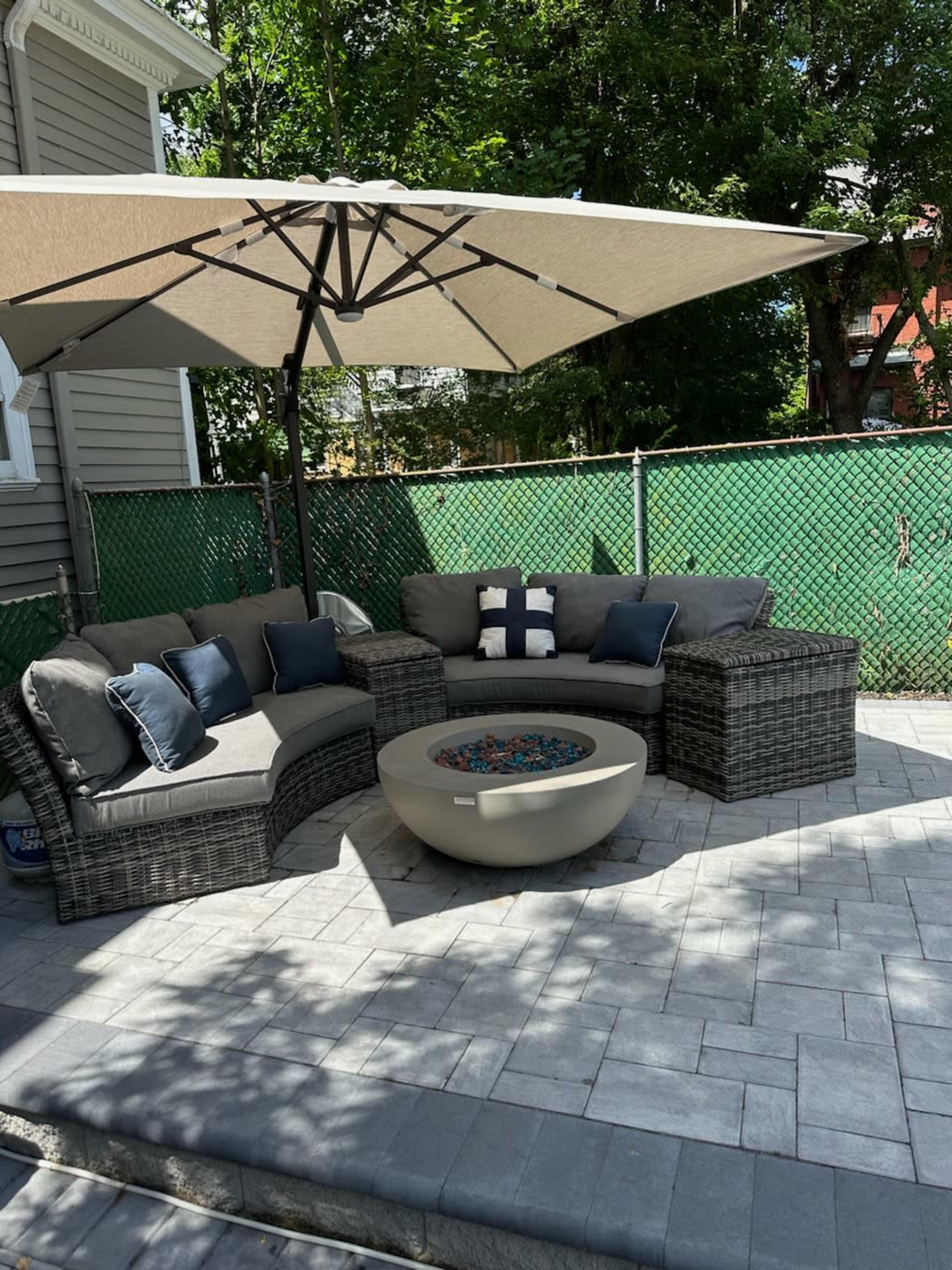A patio seating area with a sectional sofa, an umbrella, and a fire pit surrounded by a paved stone surface and a green fence in the background.
