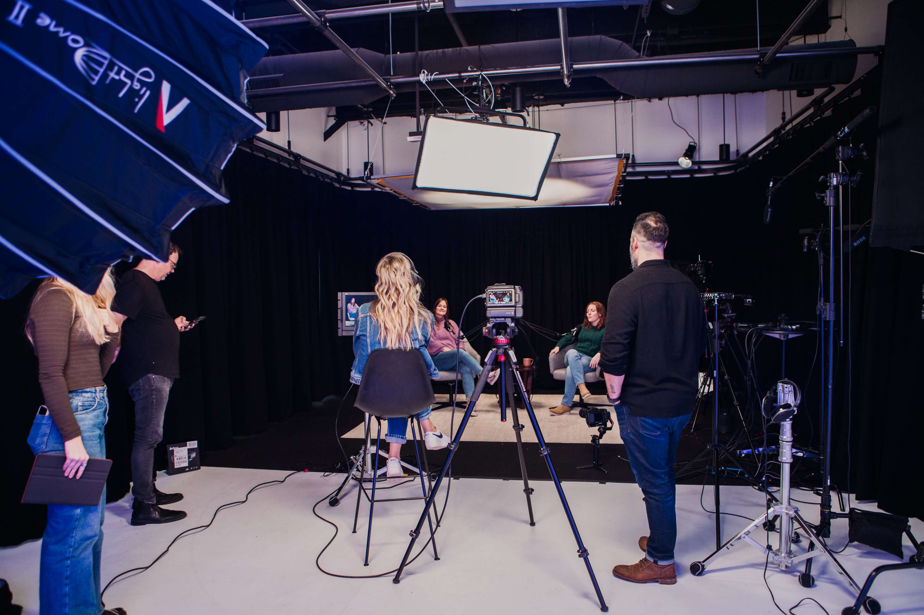 A group of people is seated in a studio setting, with cameras and lighting equipment arranged around them for a recording session.