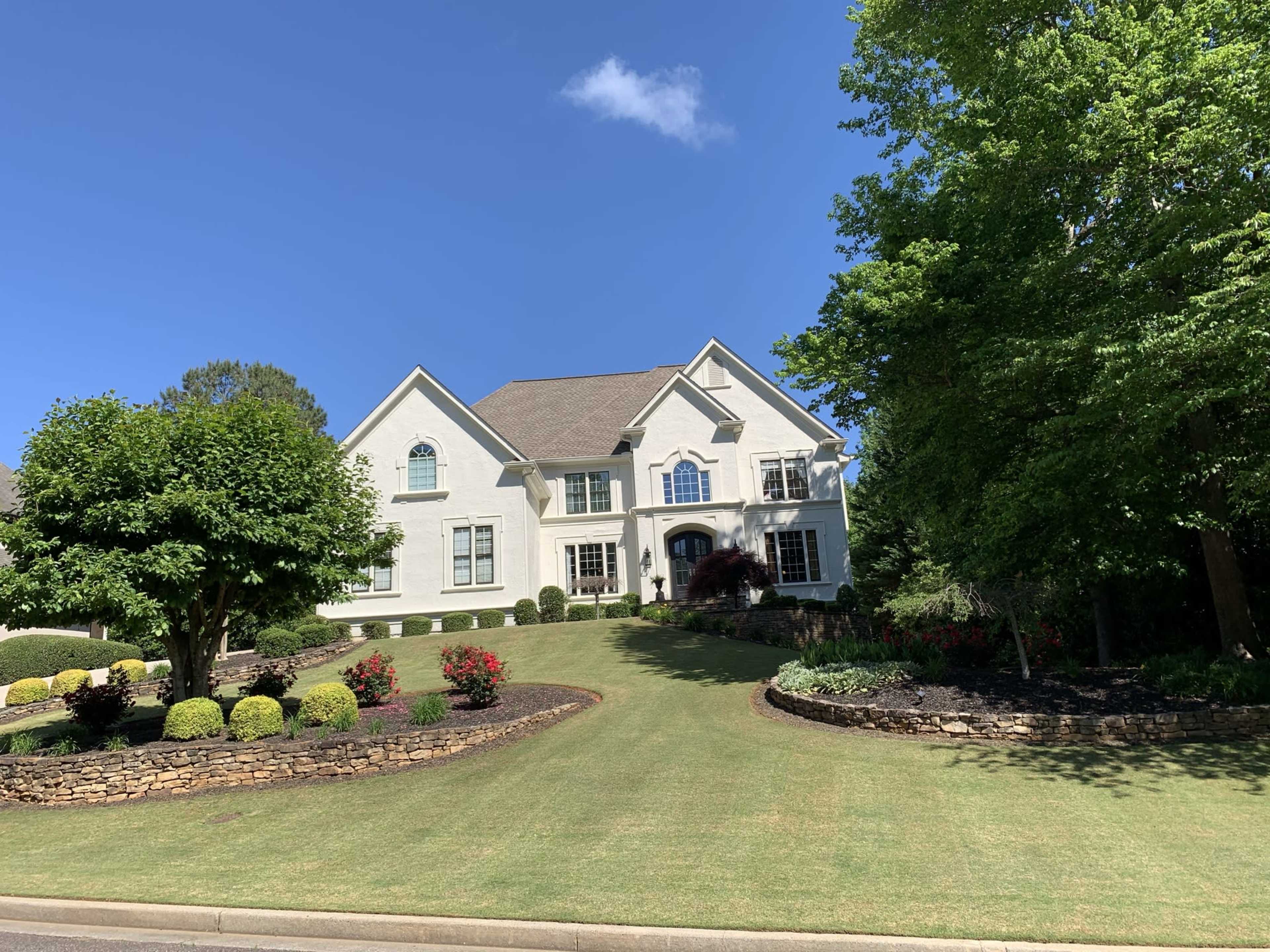 A two-story white house with a manicured lawn and landscaped gardens is set against a clear blue sky.
