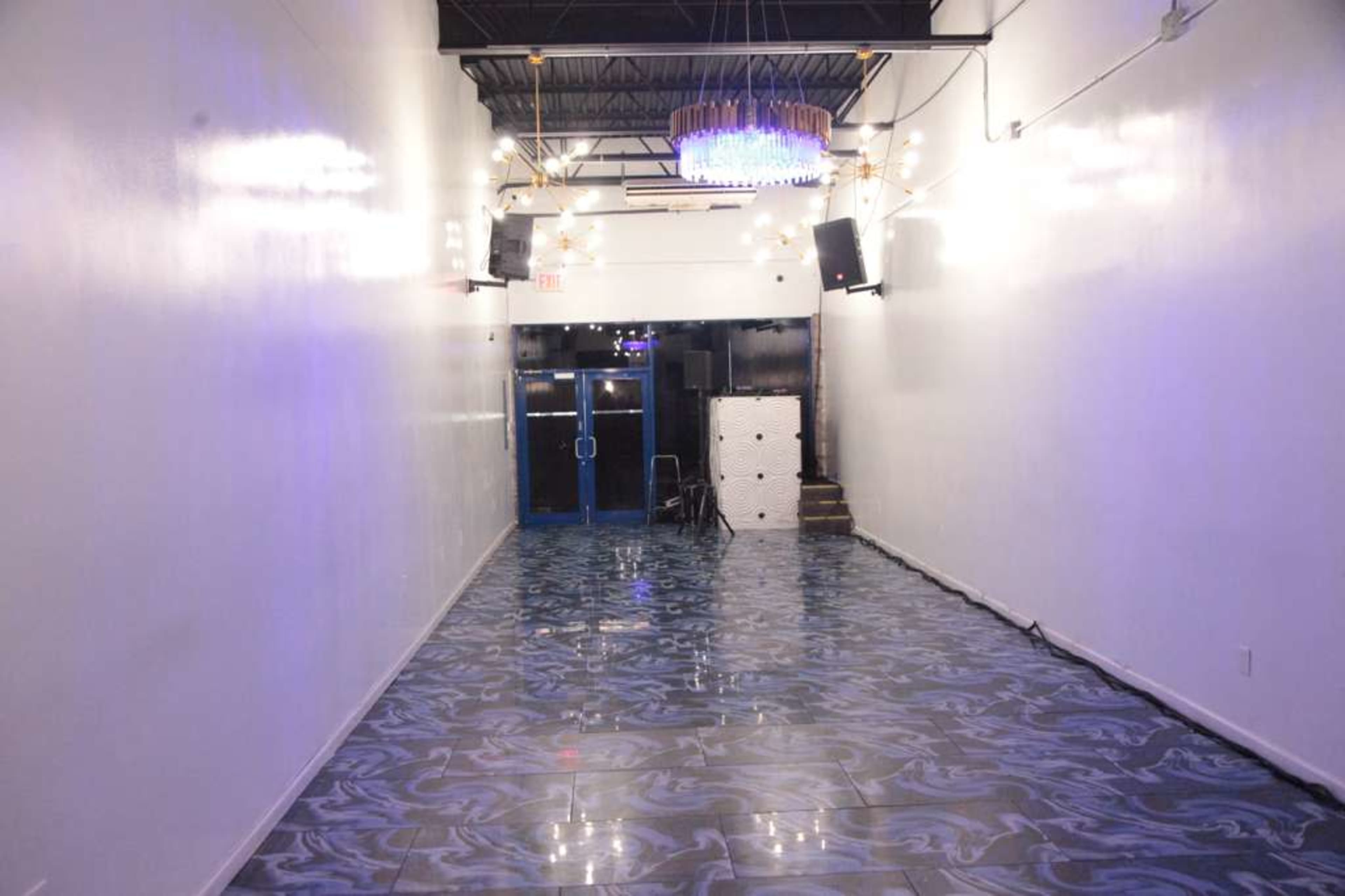 The image features a long, narrow hallway with glossy patterned flooring, illuminated by overhead lights and a chandelier, leading to double doors at the end.