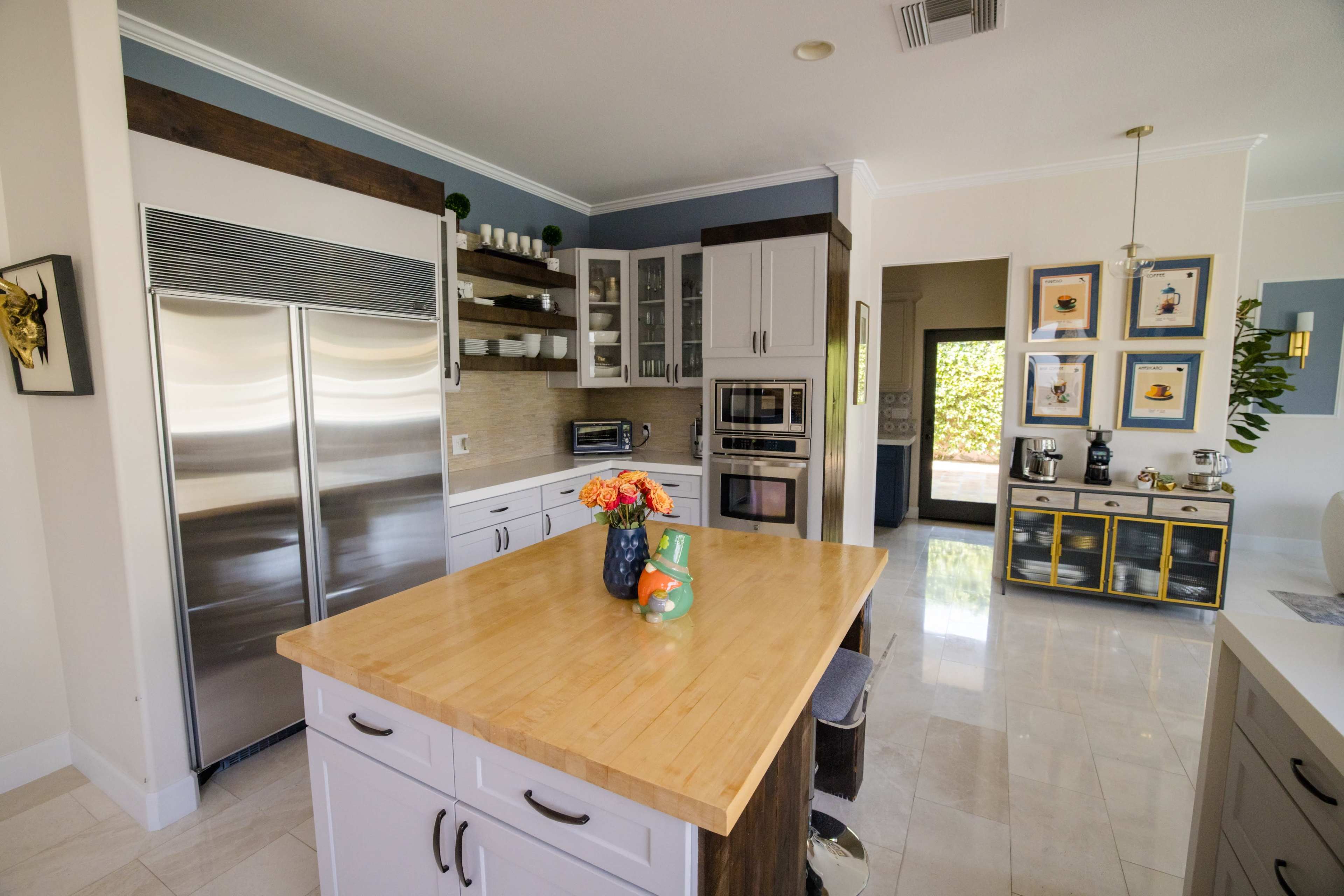 The image shows a modern kitchen featuring stainless steel appliances, wooden countertops, and decorative shelves with various items, leading to an adjoining room.