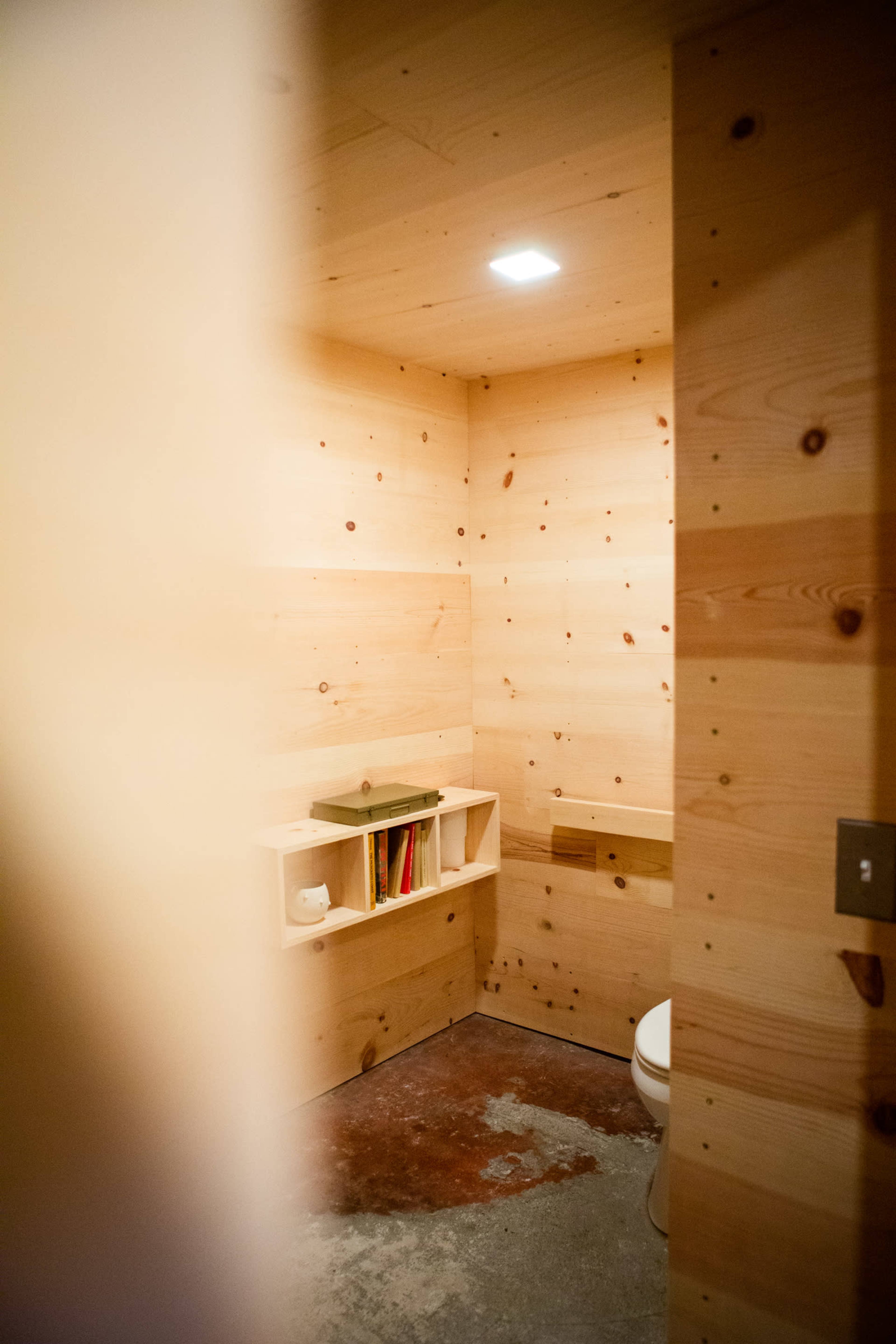 A minimalistic bathroom with wooden walls, a toilet, a small shelf containing books, and concrete flooring.