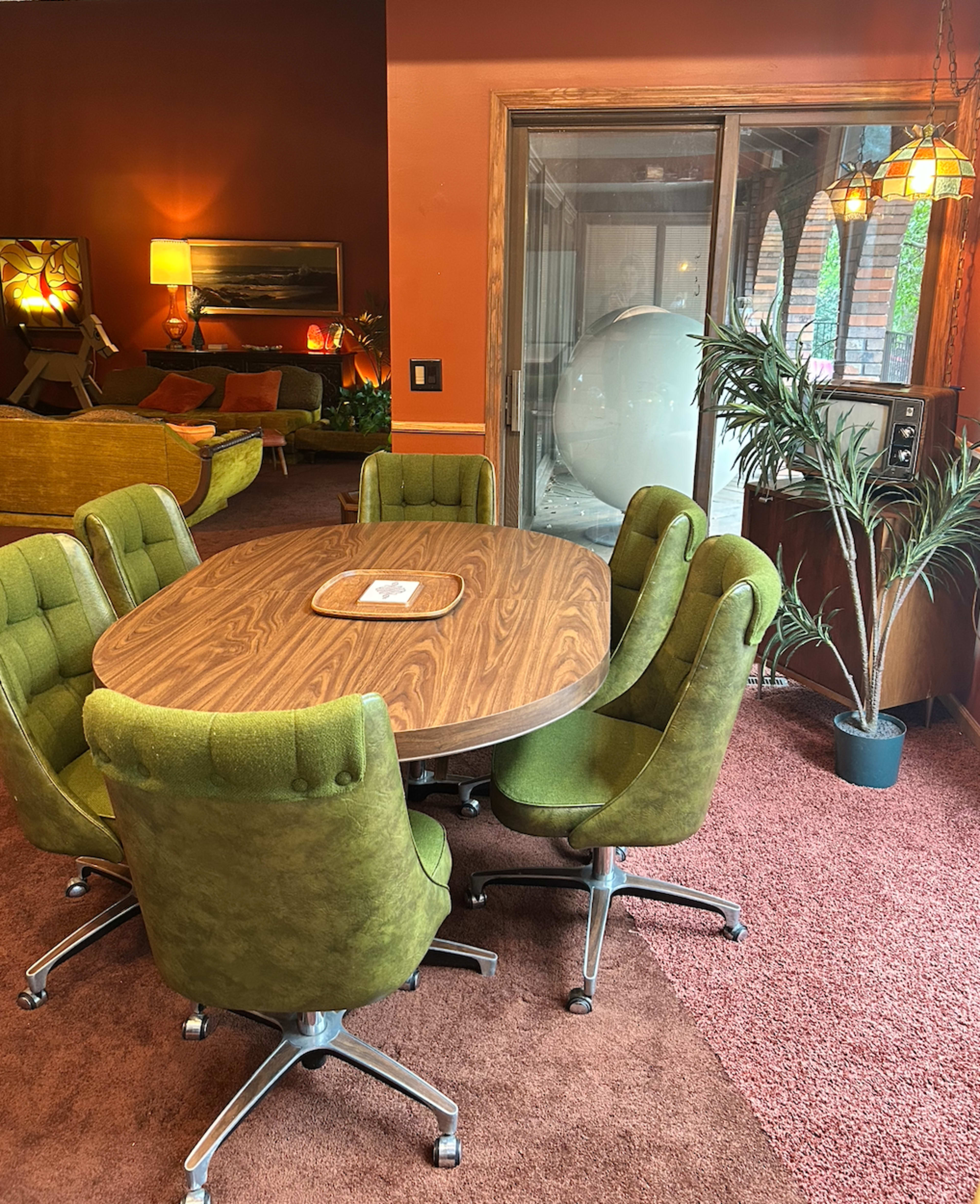 The image shows a round wooden dining table surrounded by six green upholstered chairs in a room with reddish-brown walls and ambient lighting.