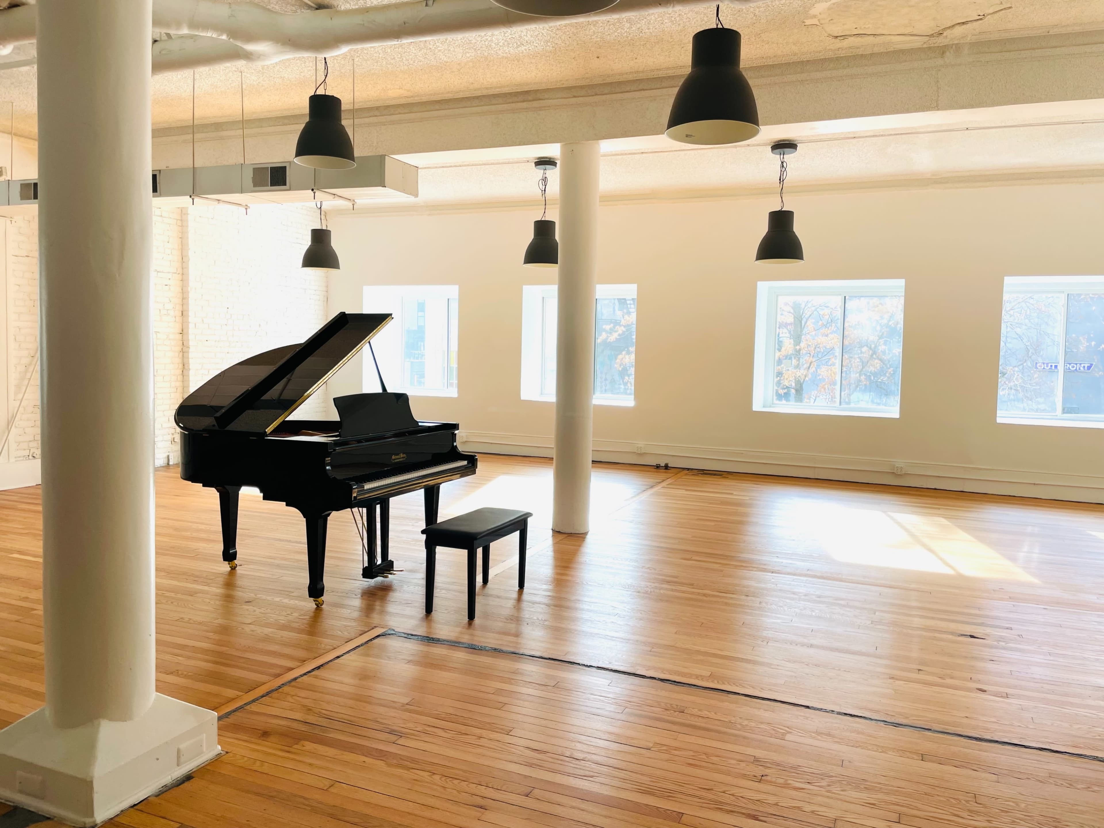 A grand piano with a bench is positioned in the center of a spacious, well-lit room with hardwood floors and large windows.