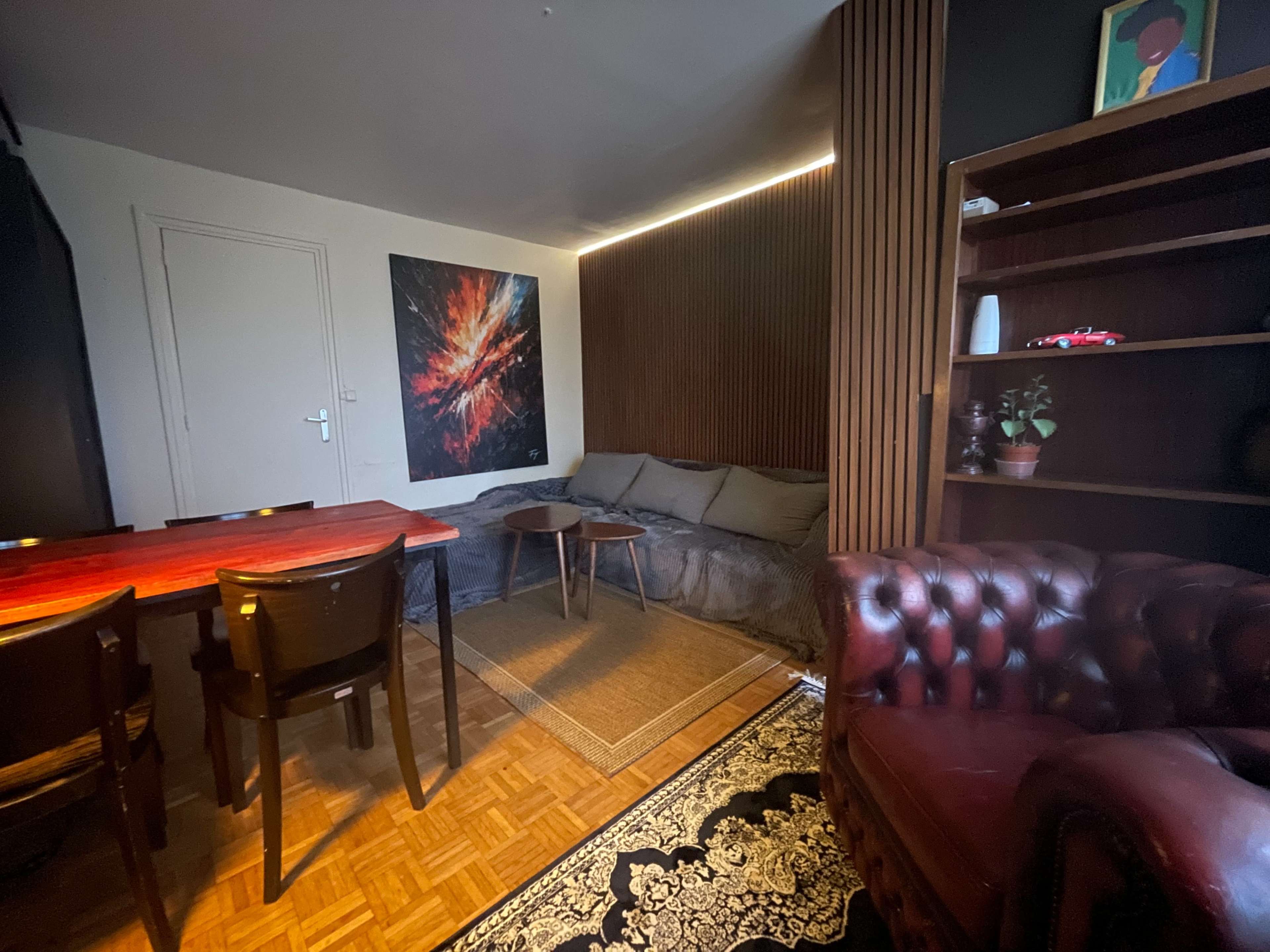 The image shows a cozy living room with a dark sofa, a wooden table with chairs, a decorative rug, and wooden paneling on one wall.
