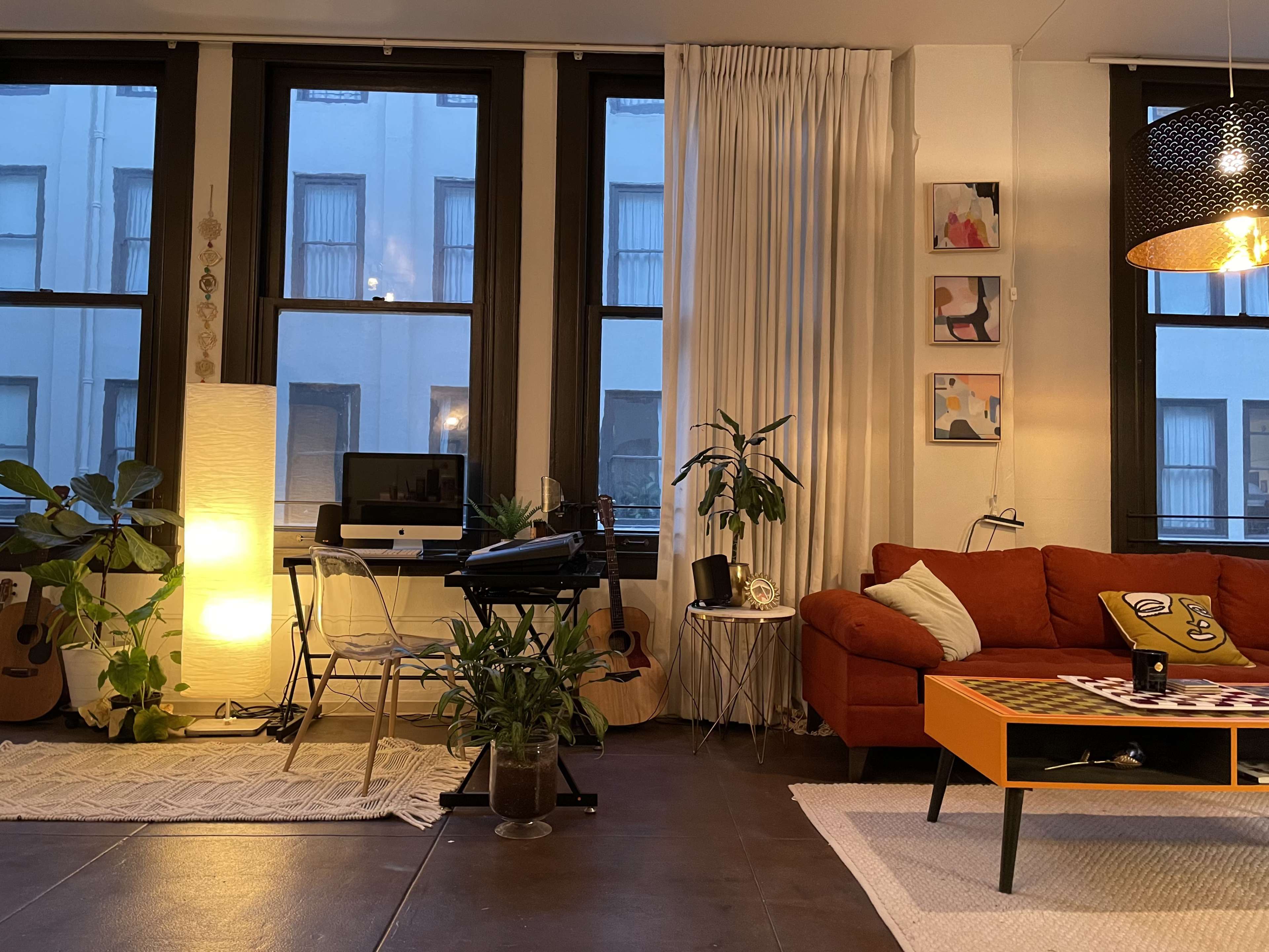 A cozy living room with a computer setup, orange couch, plants, and artwork on the walls.