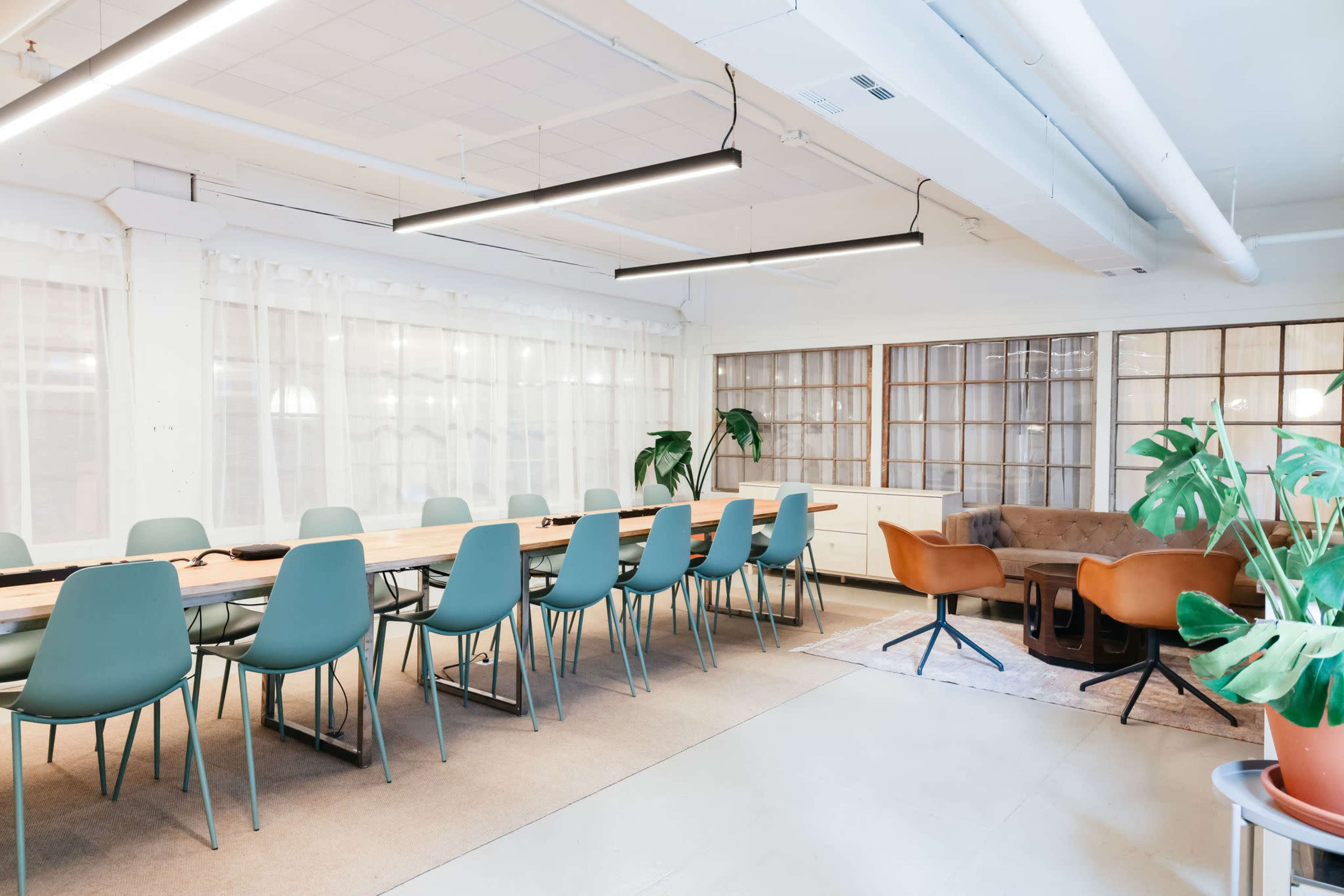A modern meeting room with a long table surrounded by green chairs, a brown sofa, and large windows covered with sheer curtains.