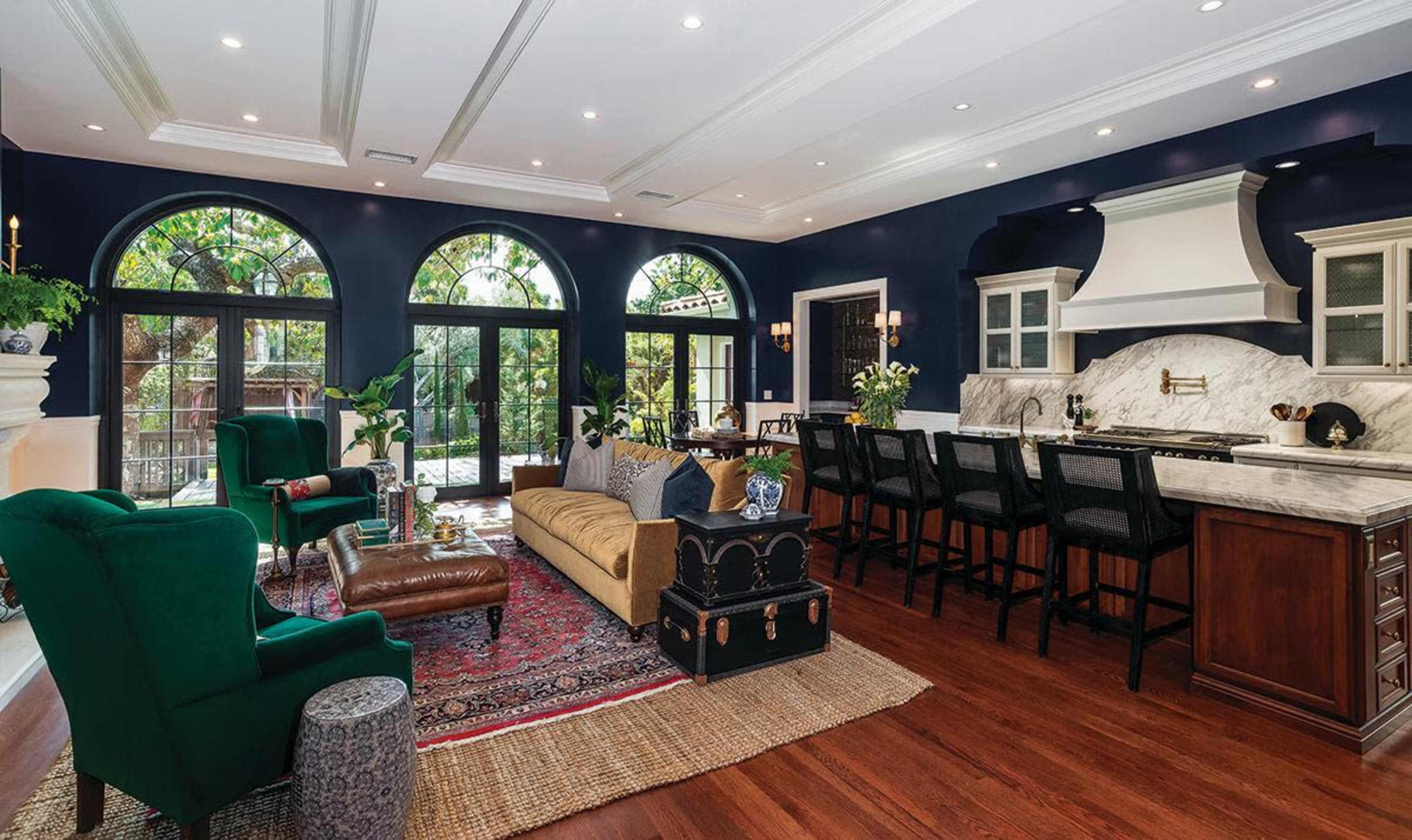 A spacious living area features a dark blue color scheme, large arched windows, and a mix of elegant furniture including a green velvet chair and a patterned area rug.