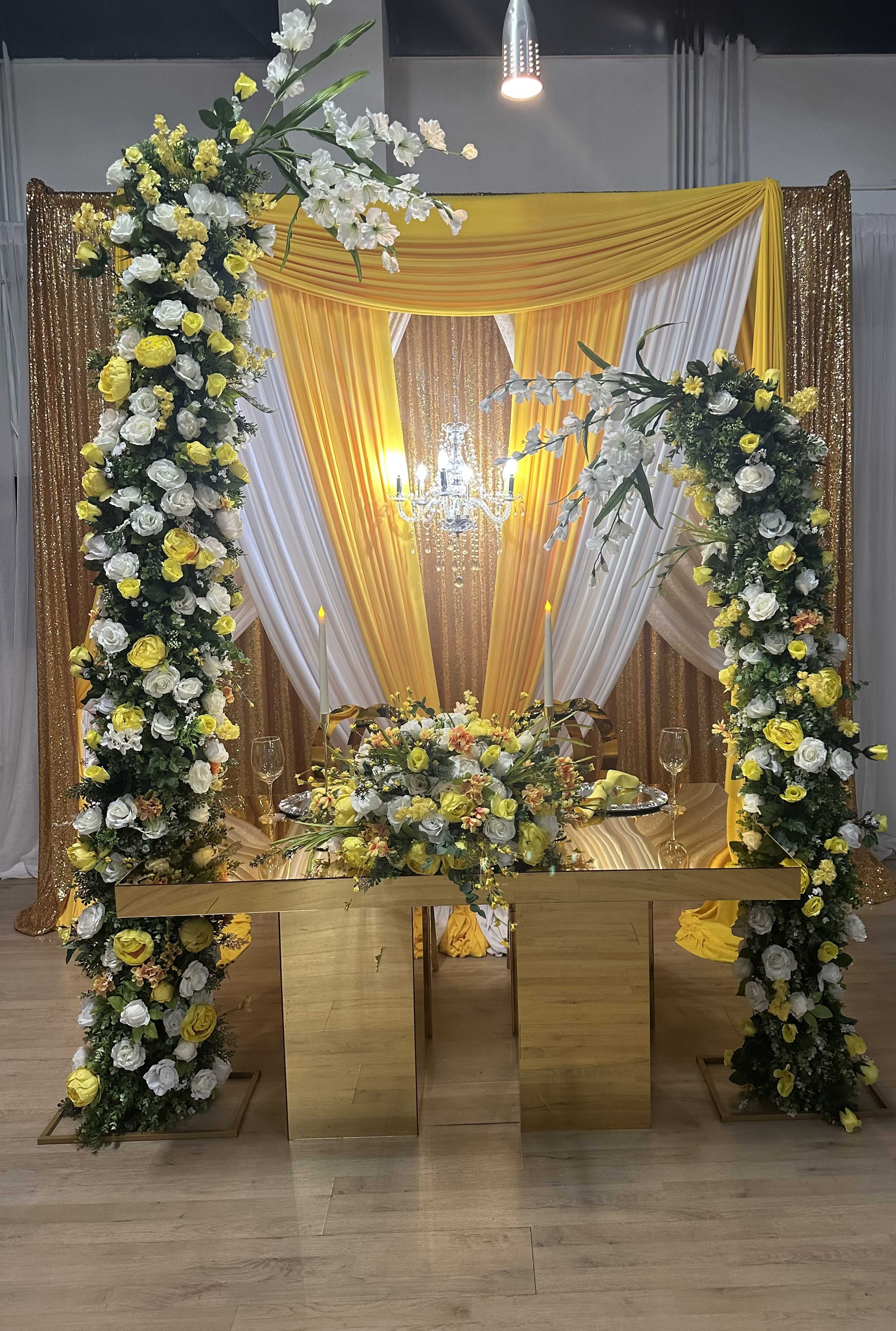 A decorated event space featuring a gold sequined backdrop, floral arrangements in yellow and white, and a table set for a celebration.