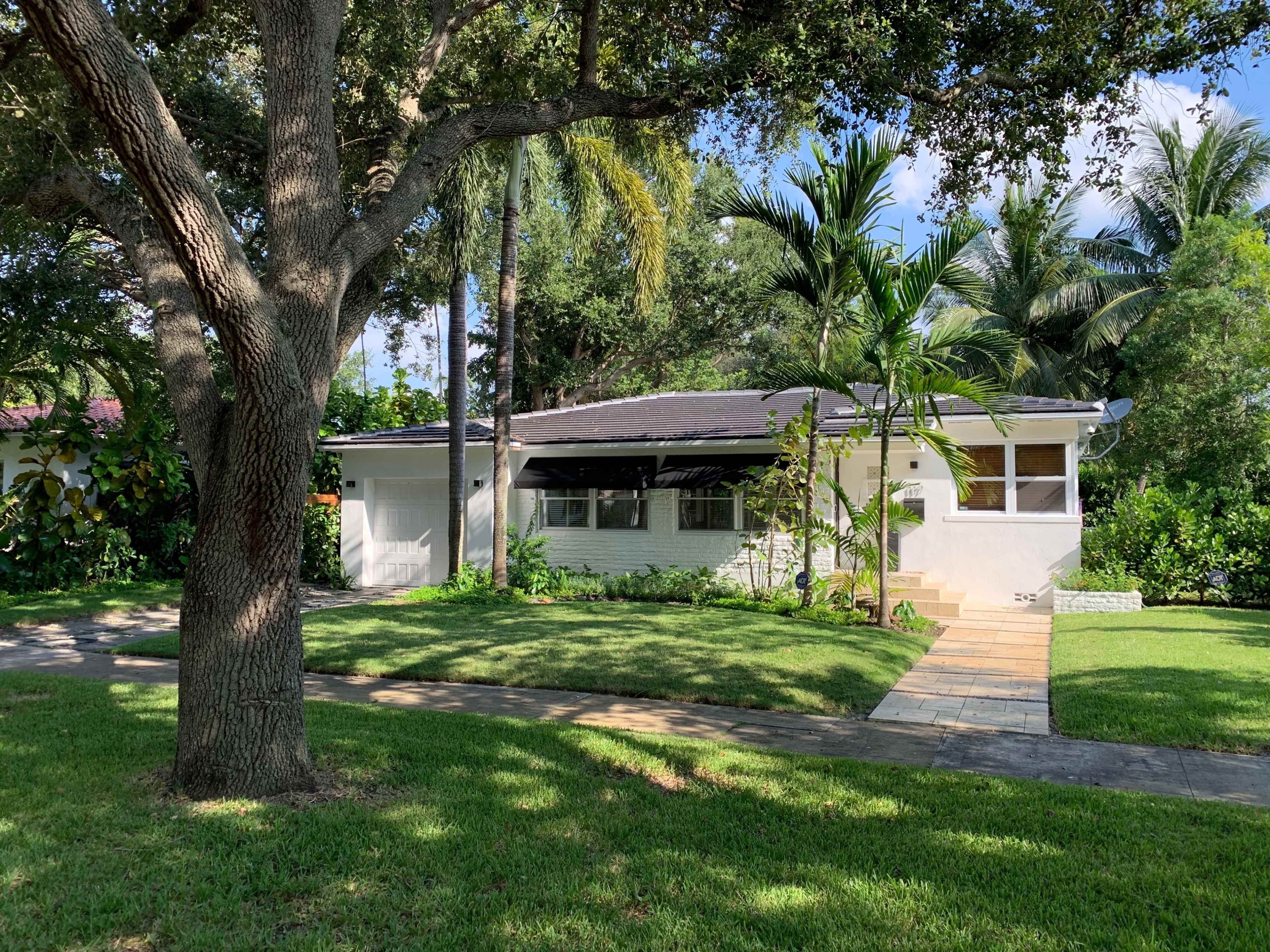A single-story house with a front porch is surrounded by green grass and tropical plants under a clear blue sky.