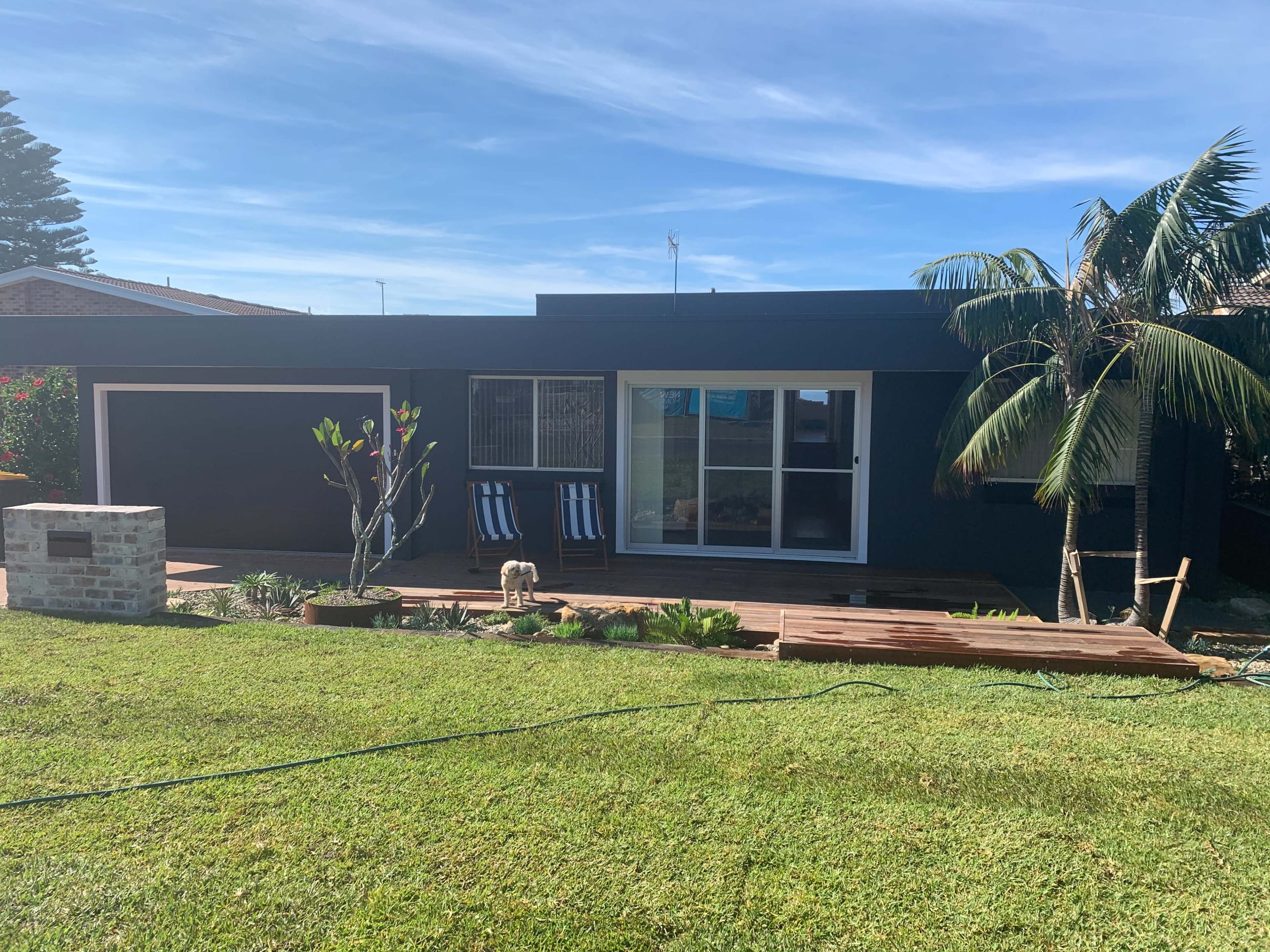 A modern black house with a wooden deck, large windows, and palm trees is set against a green lawn.