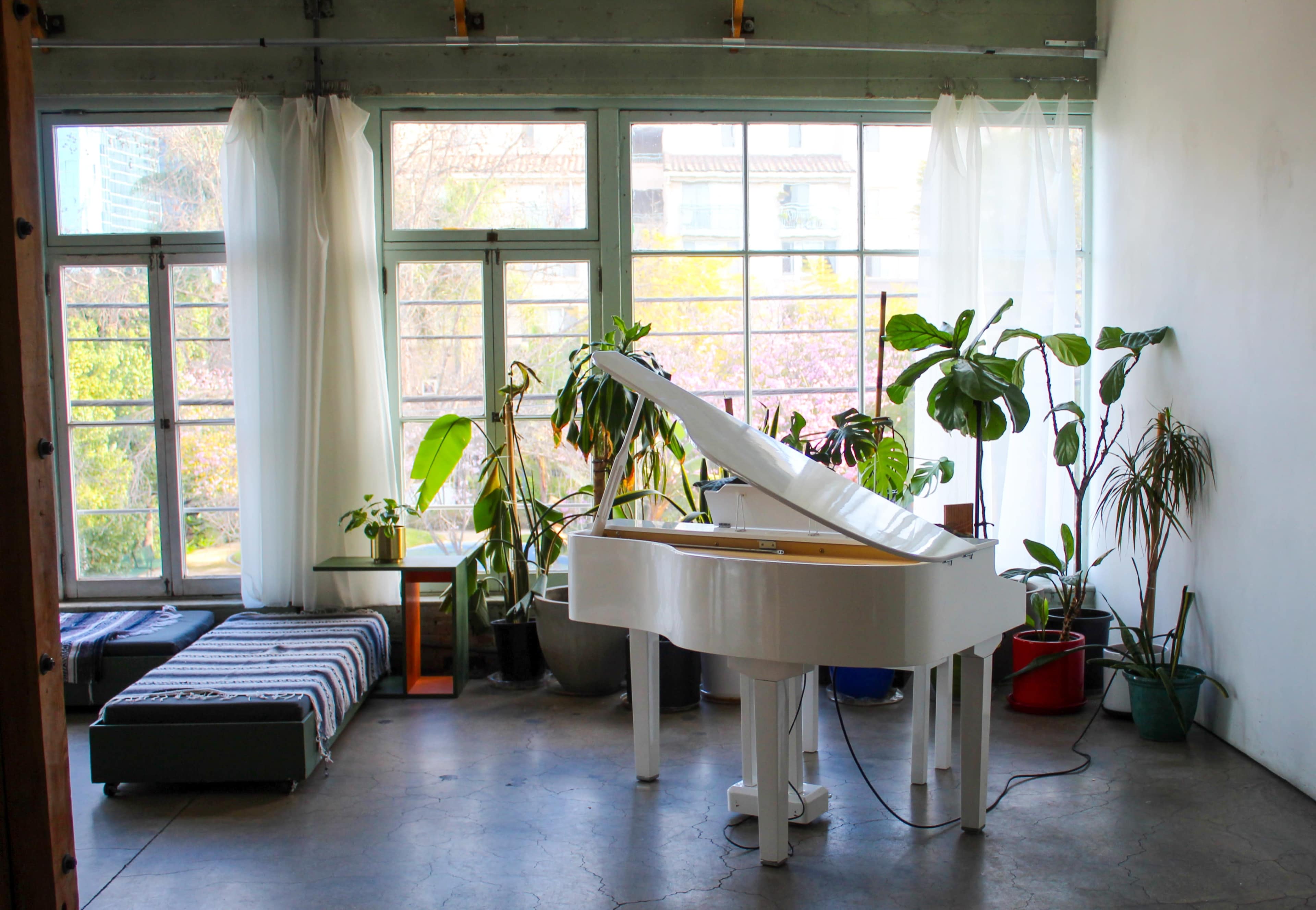 A white grand piano is positioned in a bright room filled with large windows and various potted plants.