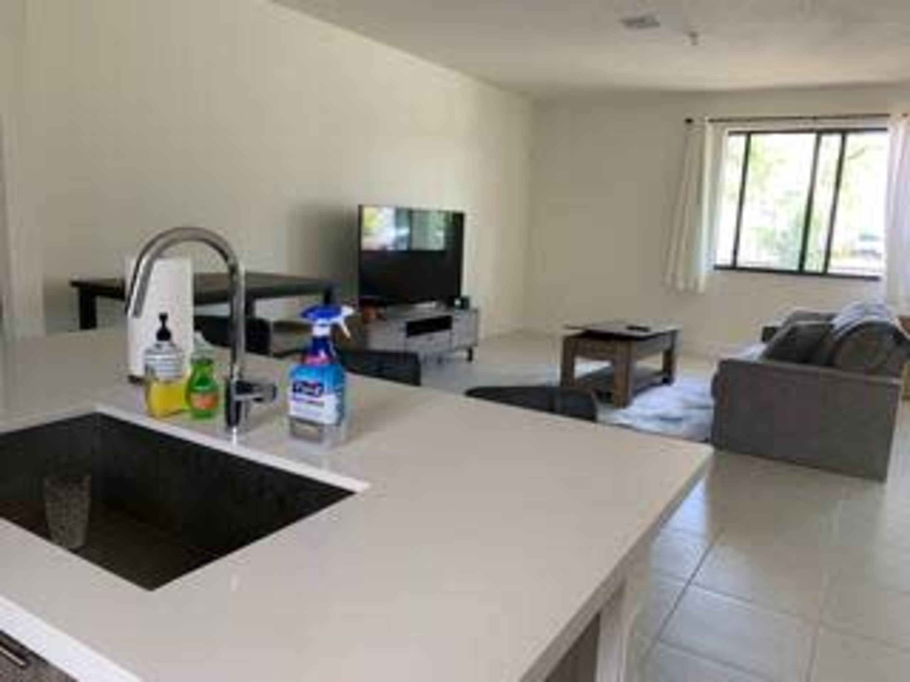 The image shows a modern living room viewed from a kitchen, featuring a large window, a television, a gray sofa, and a coffee table.