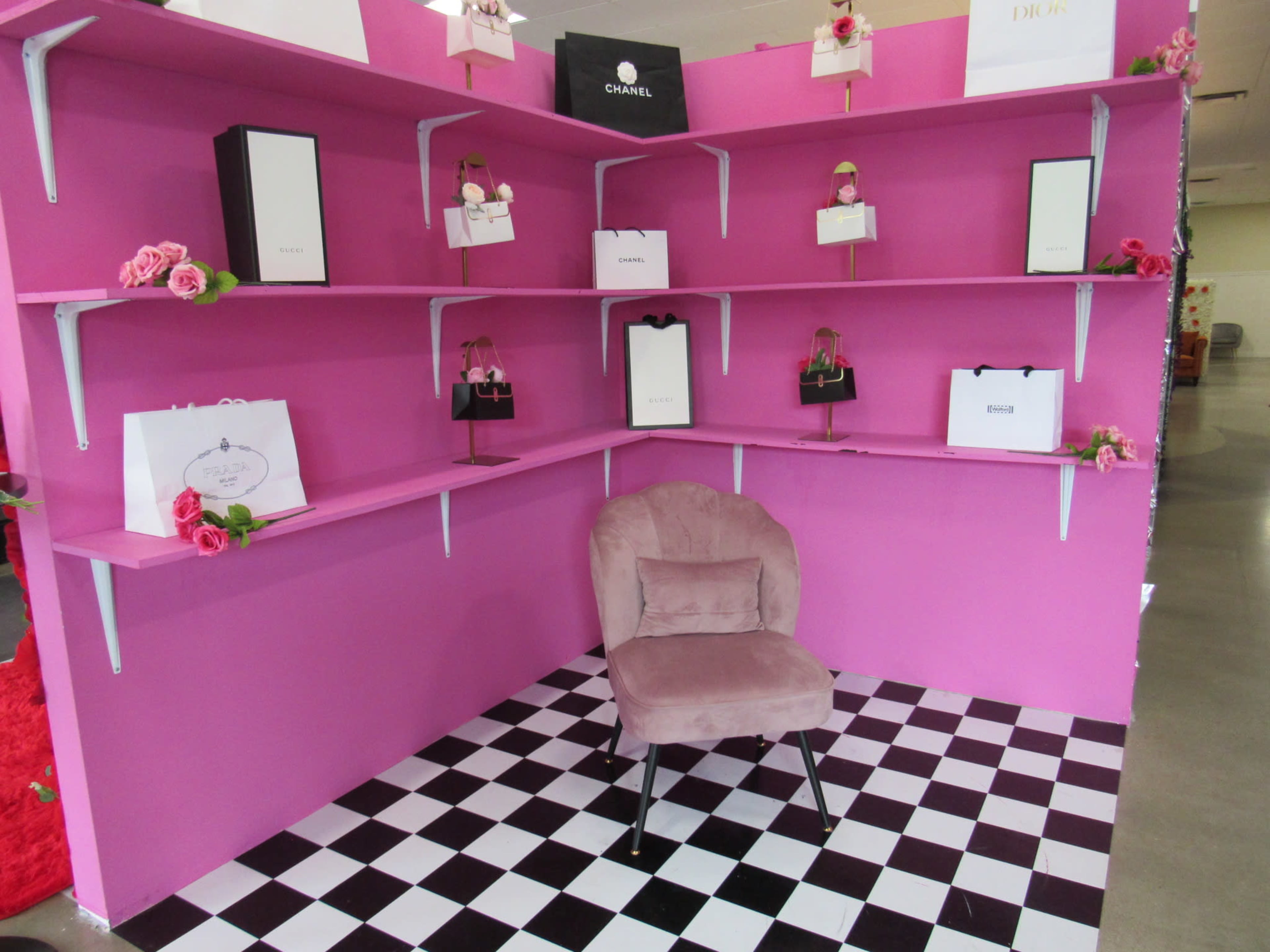 A pink wall with white shelves displays various branded shopping bags and boxes, alongside a single plush chair on a black-and-white checkered floor.