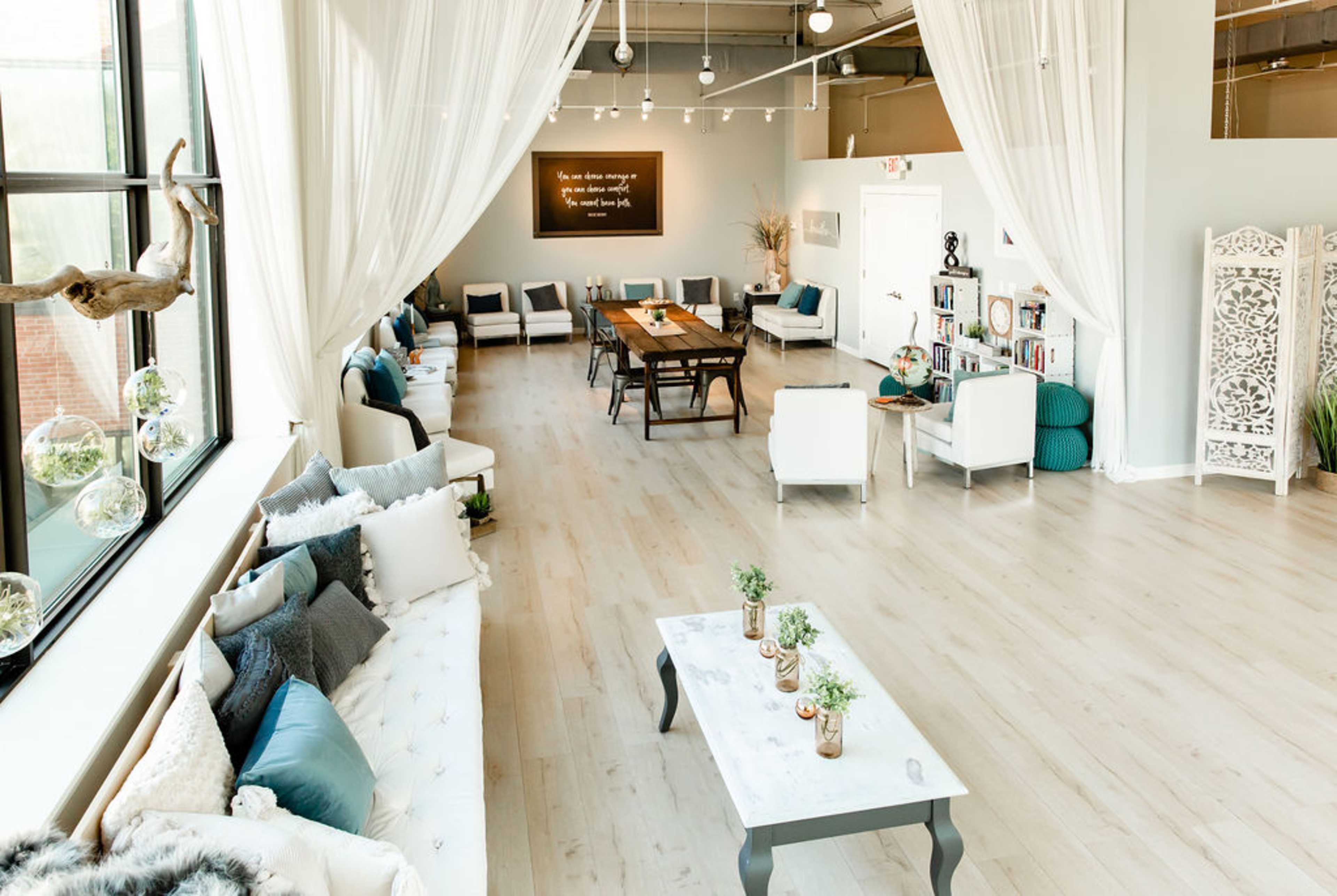 A spacious, well-lit room features a long wooden table surrounded by seating, with white drapes and plants decorating the space.
