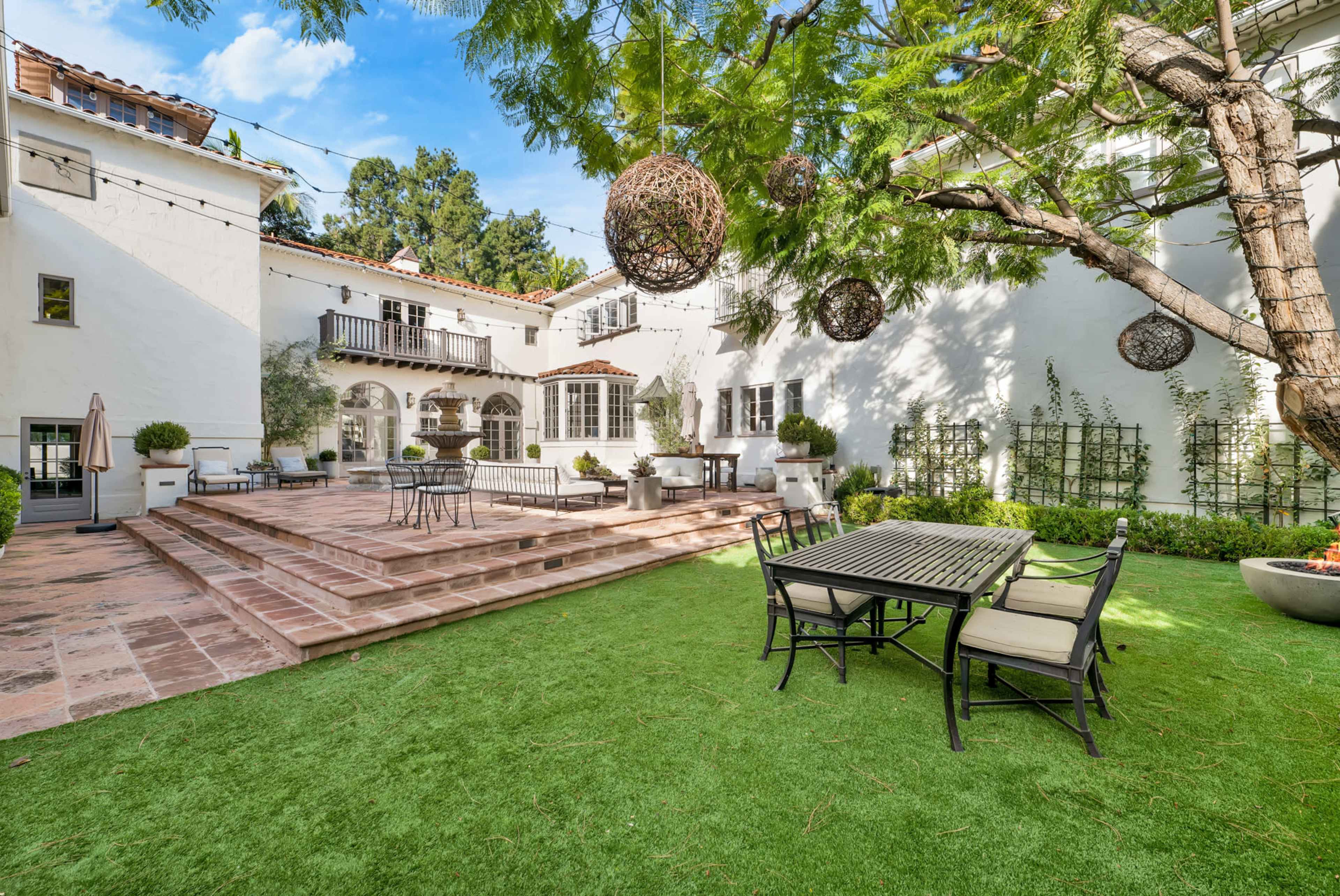 A landscaped outdoor patio area featuring a stone terrace, dining table, and a large tree with hanging decorations.