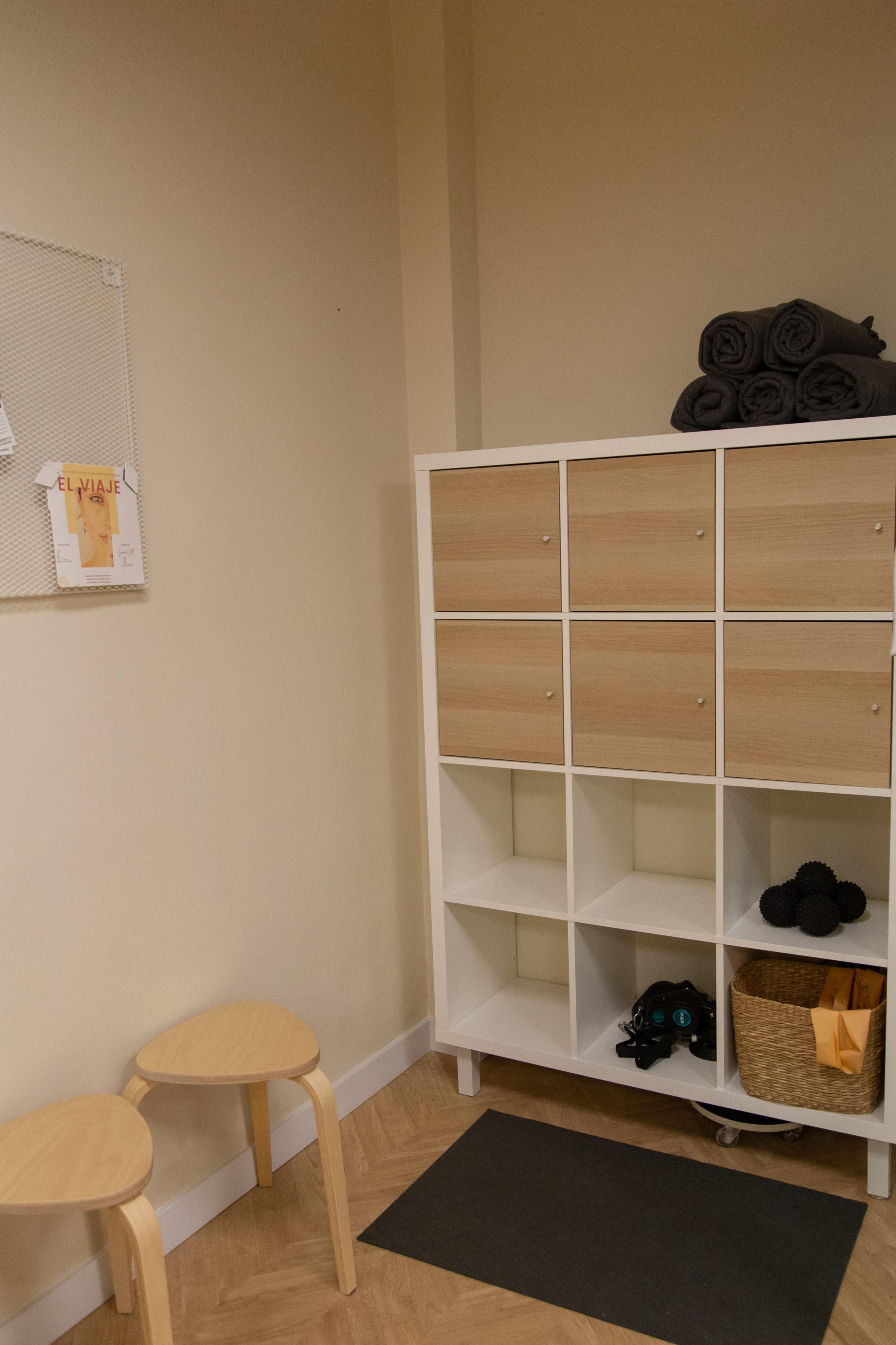 A small room with a white storage unit featuring cubbies and a basket, two wooden stools, and a wall with a notice board.