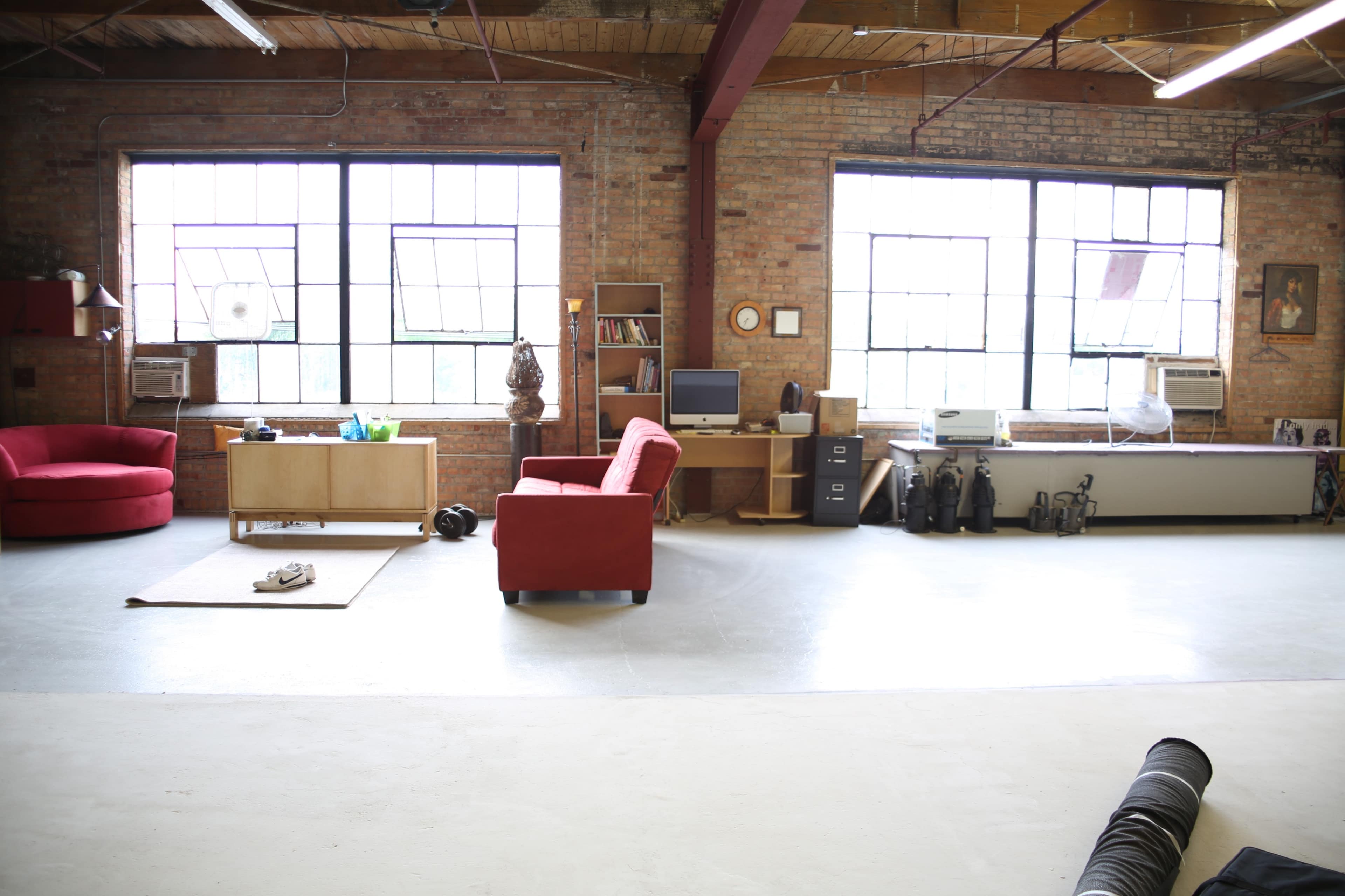 A spacious industrial-style room with large windows, featuring a red couch, a wooden cabinet, an office desk with a computer, and various equipment along the wall.
