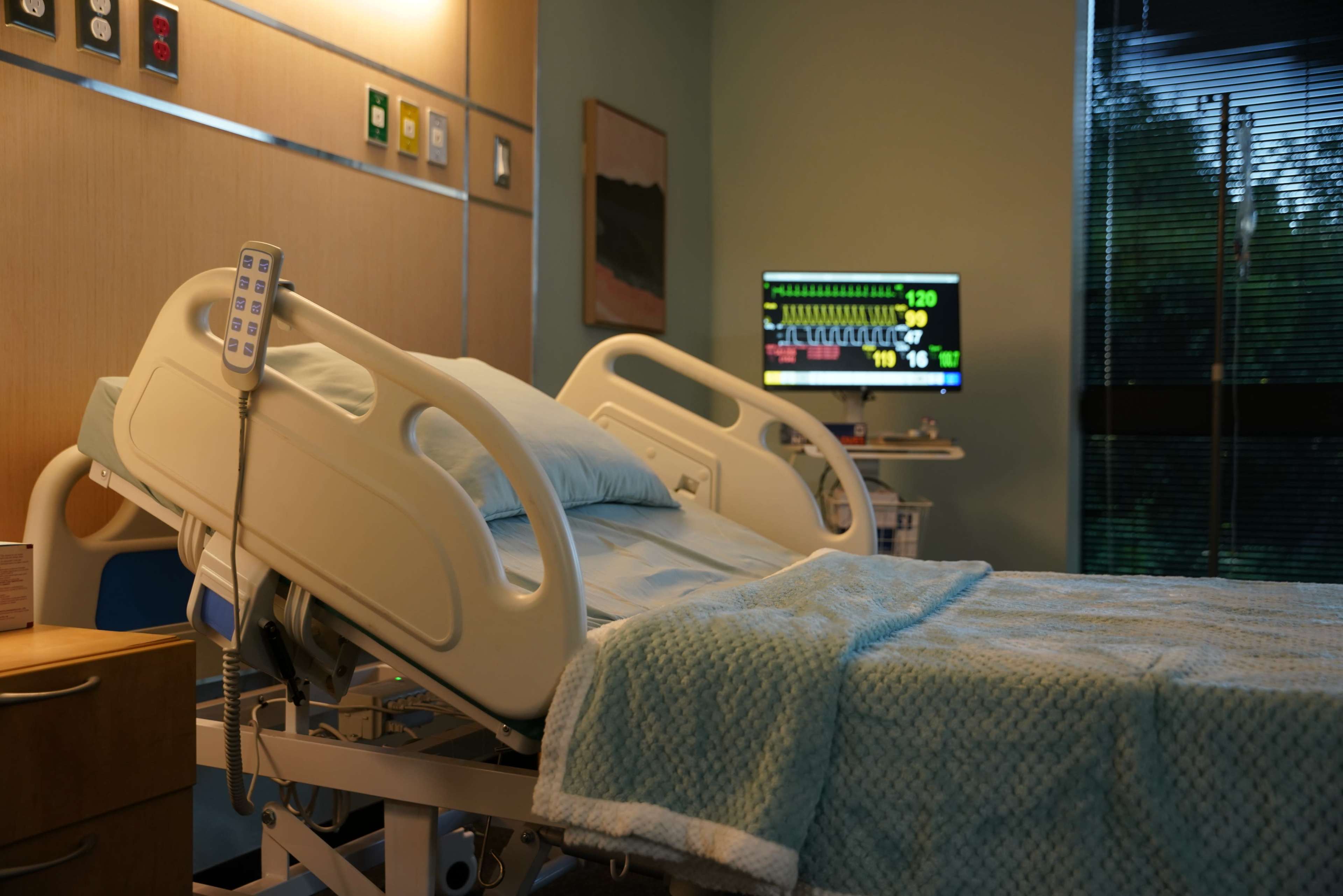 A hospital room features an adjustable bed with a blue blanket, a monitor displaying vital signs, and medical equipment nearby.