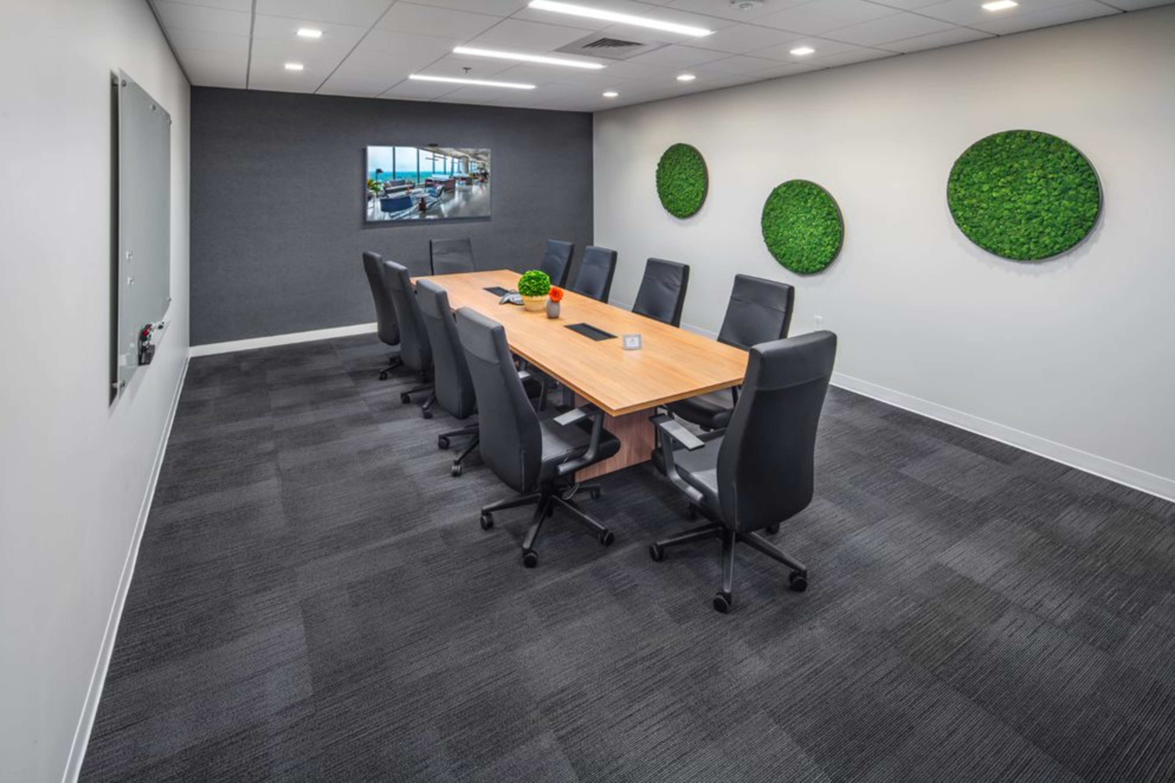 A modern conference room features a large wooden table surrounded by black chairs, with green wall accents and a screen displaying a presentation.
