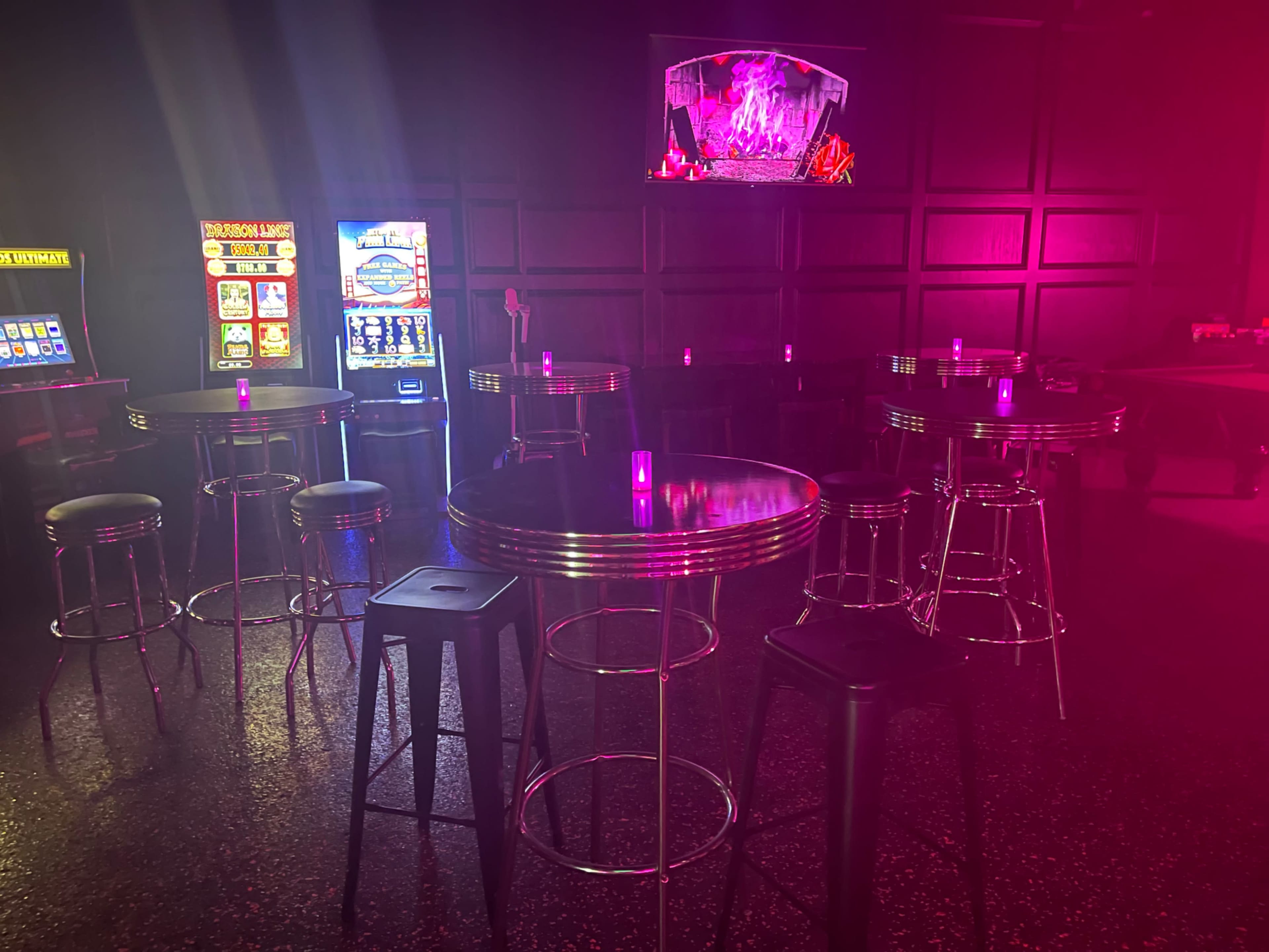 A dimly lit bar area with neon lighting, featuring several tables and chairs, along with a slot machine and a vibrant display on the wall.