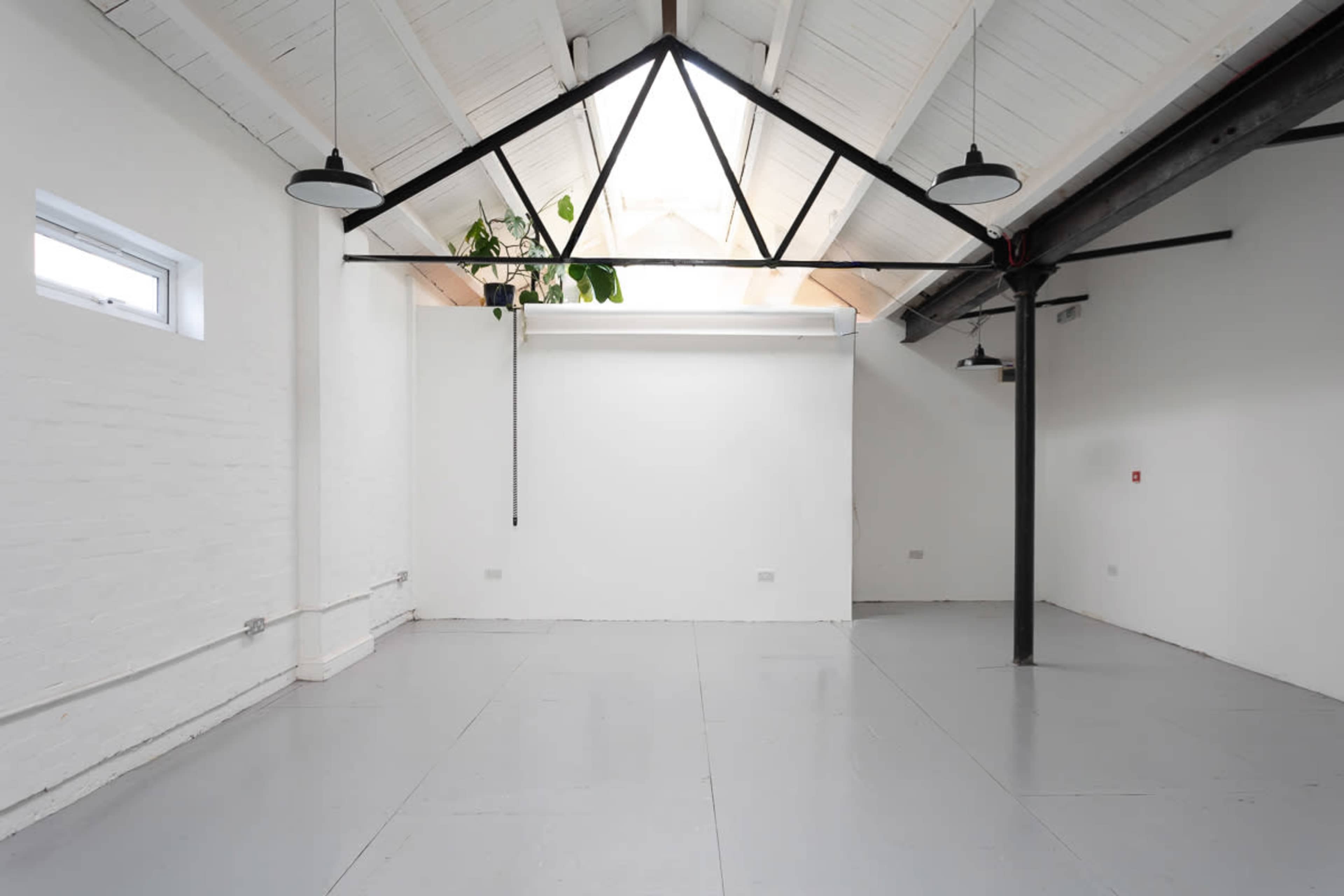 The image shows a spacious, empty room with white walls, a high ceiling, and a large skylight, featuring industrial elements like black beams and lighting.