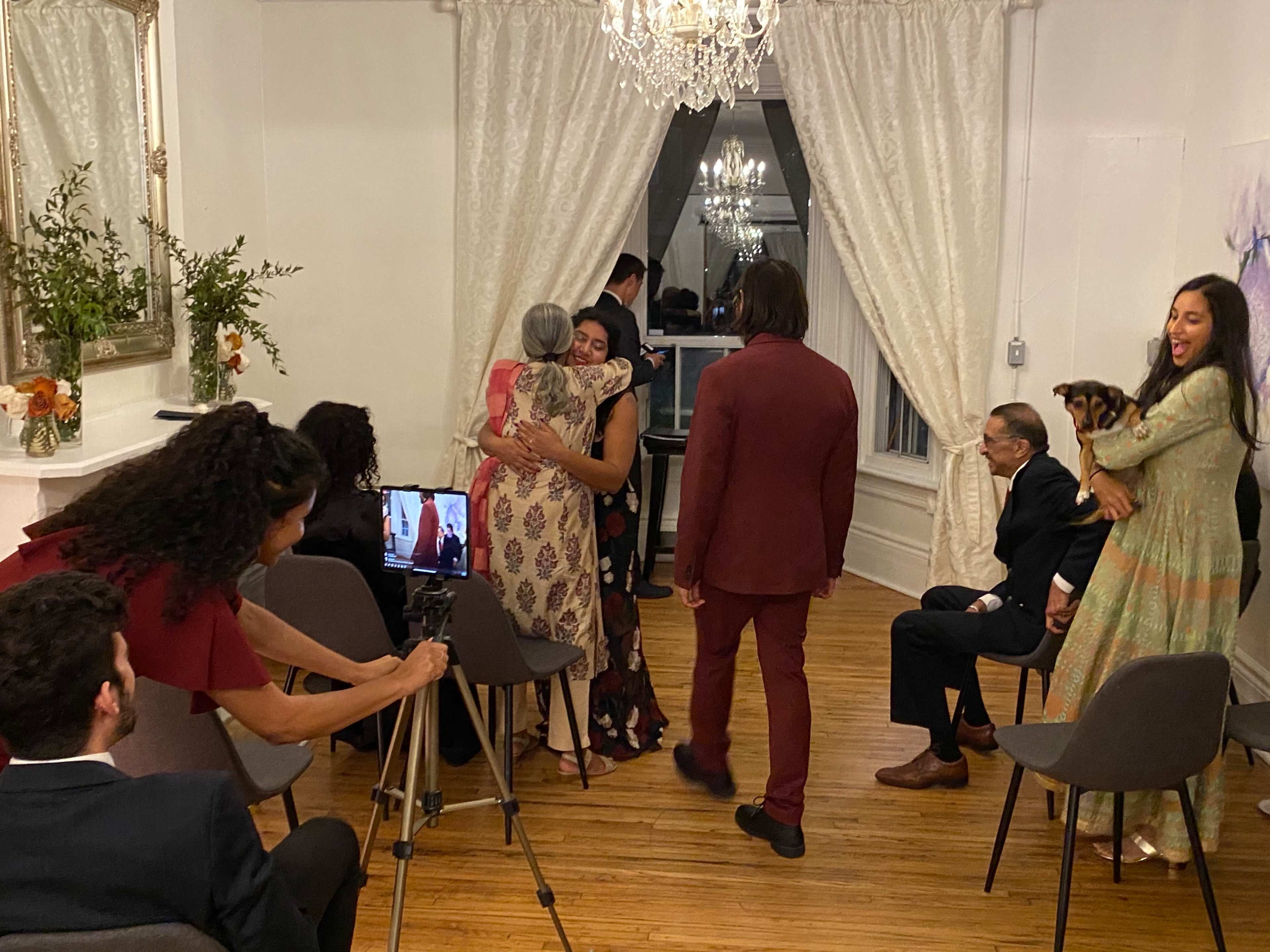 A group of people gathers in a room with a chandelier, some seated and others standing, as one woman hugs another while a camera records the moment.