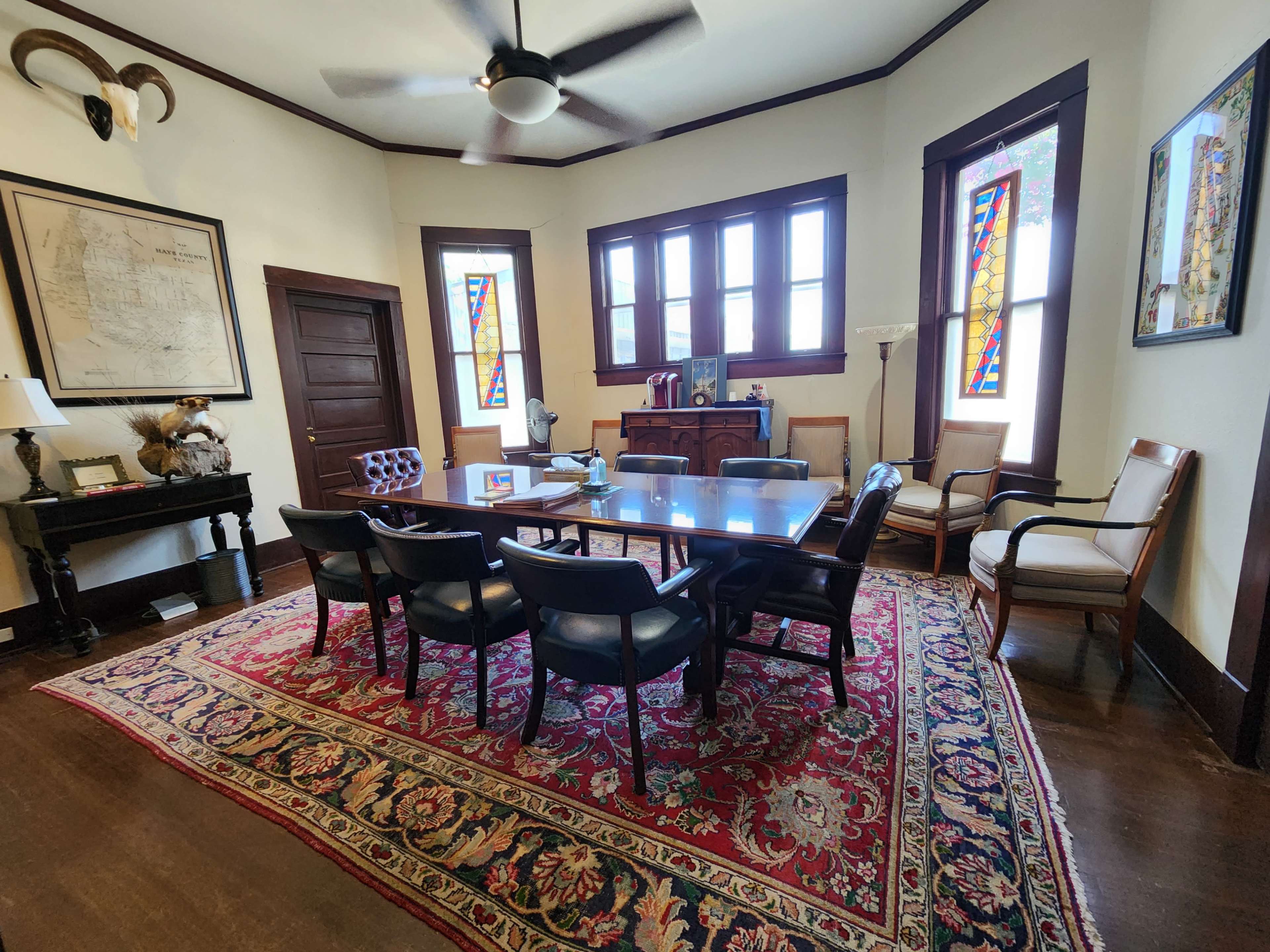 A conference room features a large wooden table surrounded by black chairs, accented by a patterned rug, with stained glass windows and various pieces of furniture along the walls.