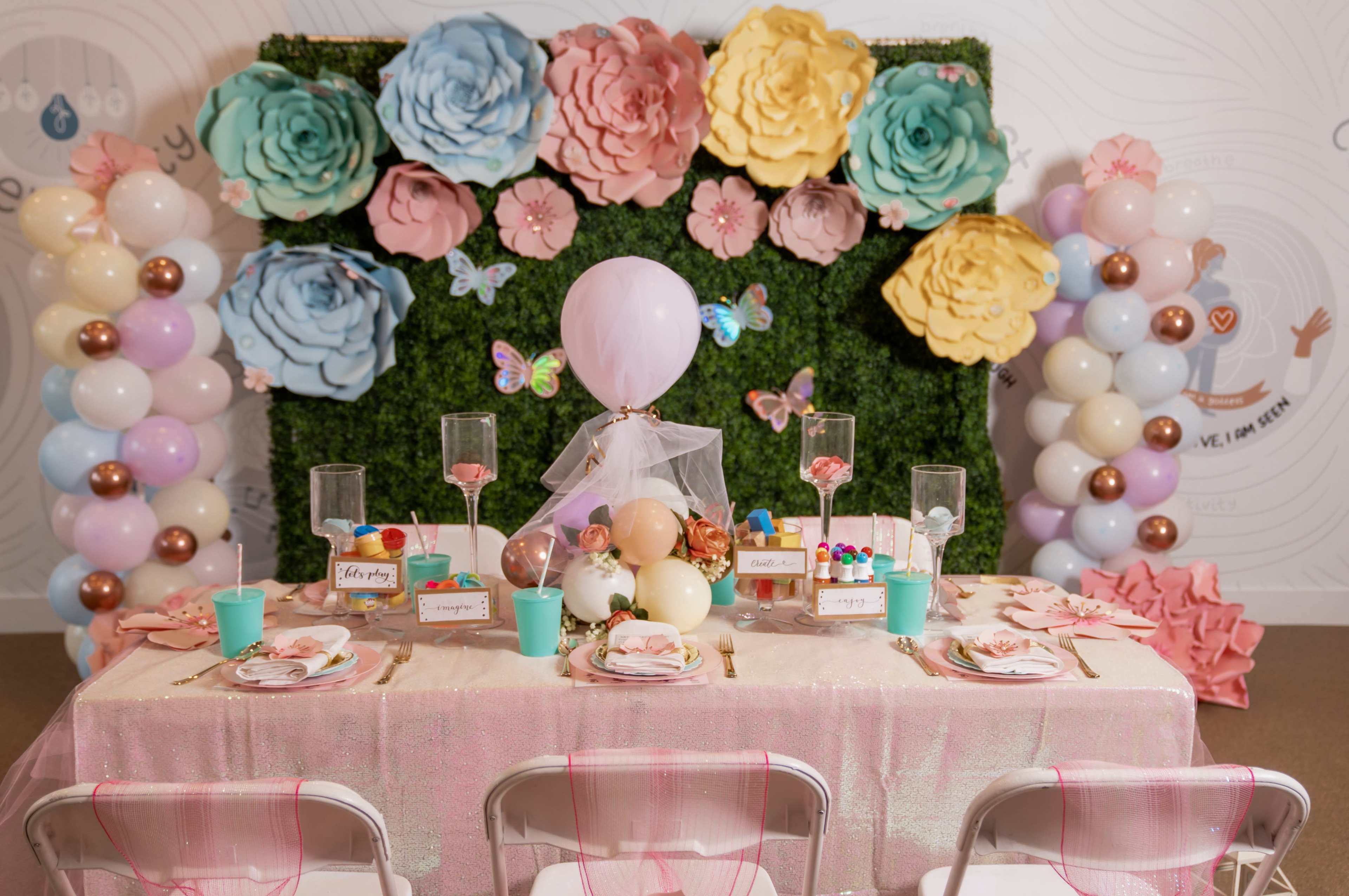 A decorated table is set against a backdrop of large paper flowers, featuring colorful balloons and a selection of desserts.