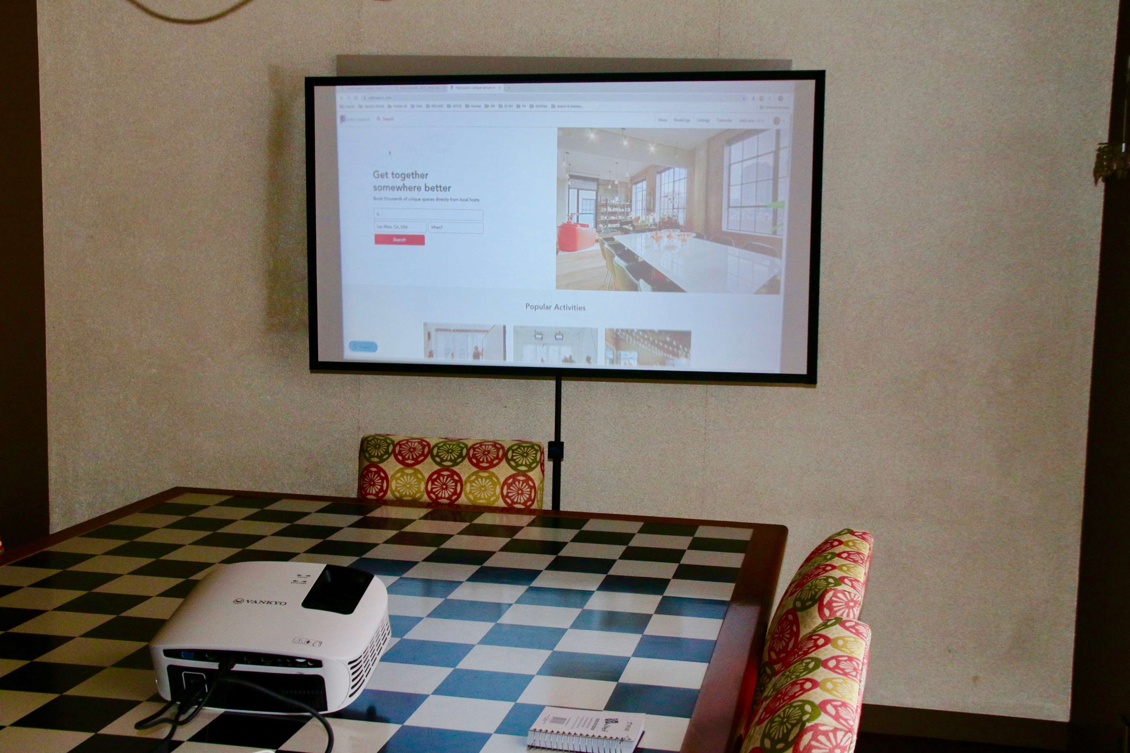 A digital projector displays a webpage on a large screen above a checkered table with colorful chairs.