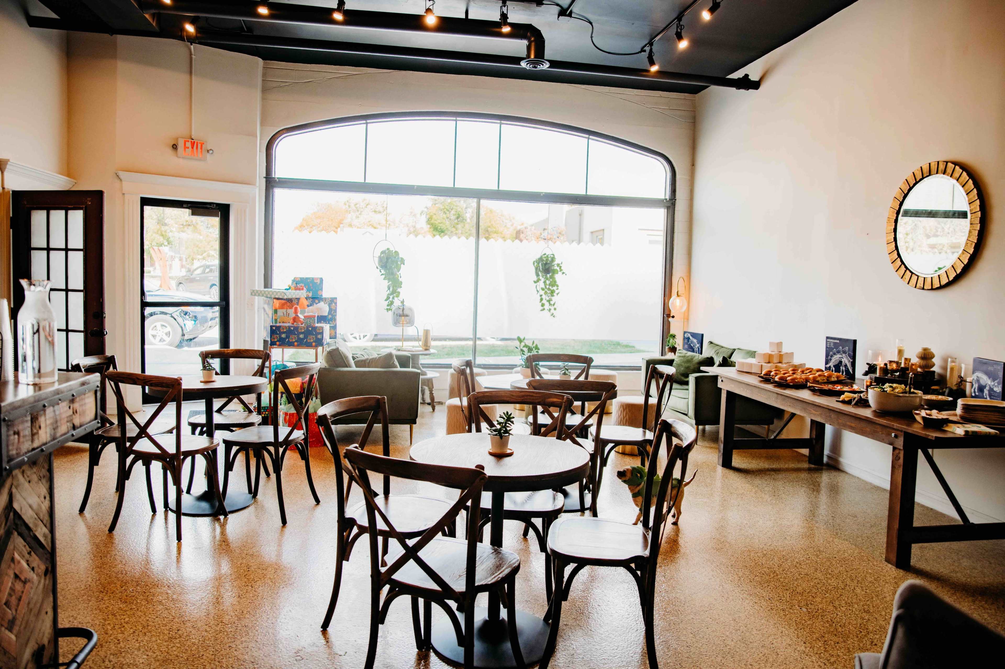 The image shows a cozy café interior with several wooden tables and chairs, a large window, and decorative plants.