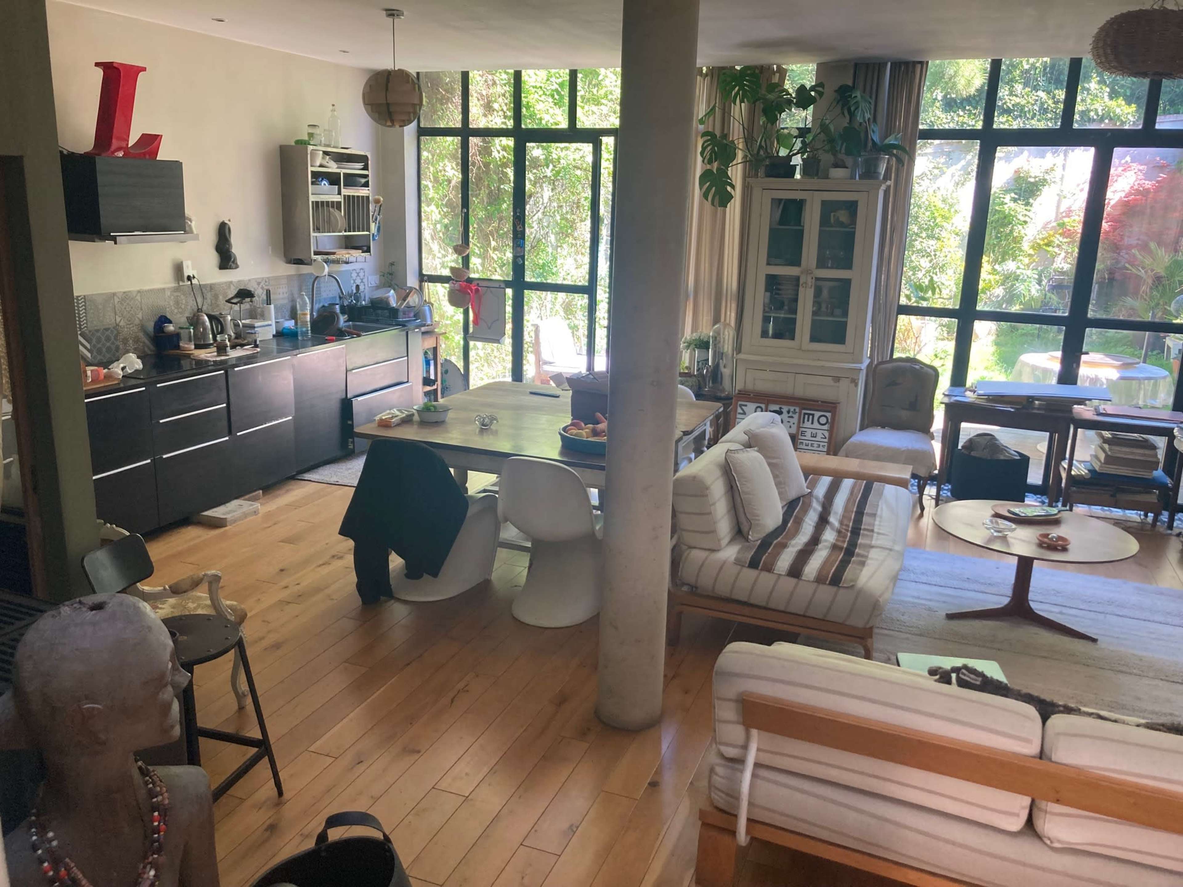 The image shows a modern living space with a kitchen, dining area, and large windows overlooking a garden.