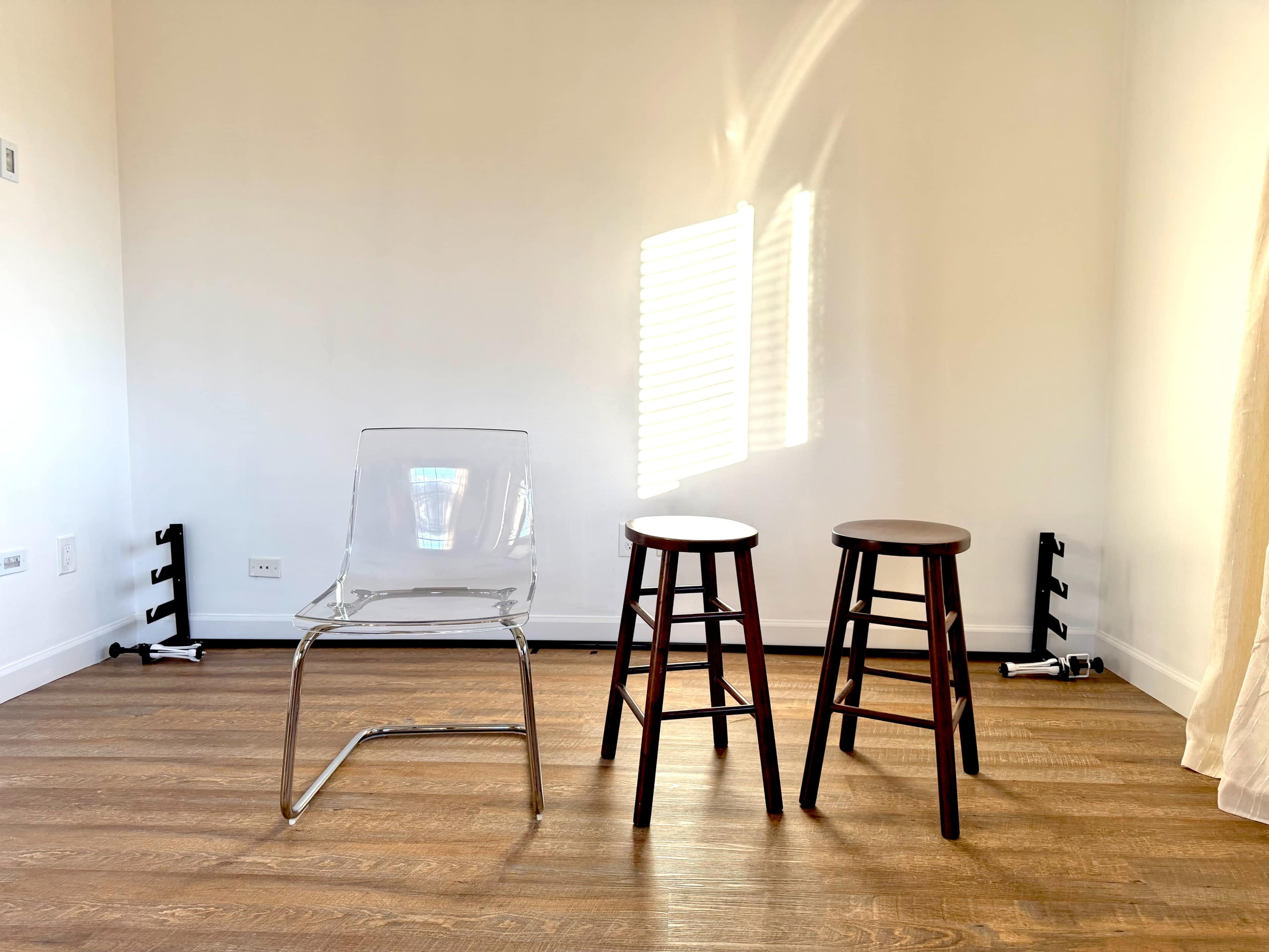 The image shows a minimalist room with a clear chair and two wooden stools arranged on a wooden floor, with sunlight streaming through a window.