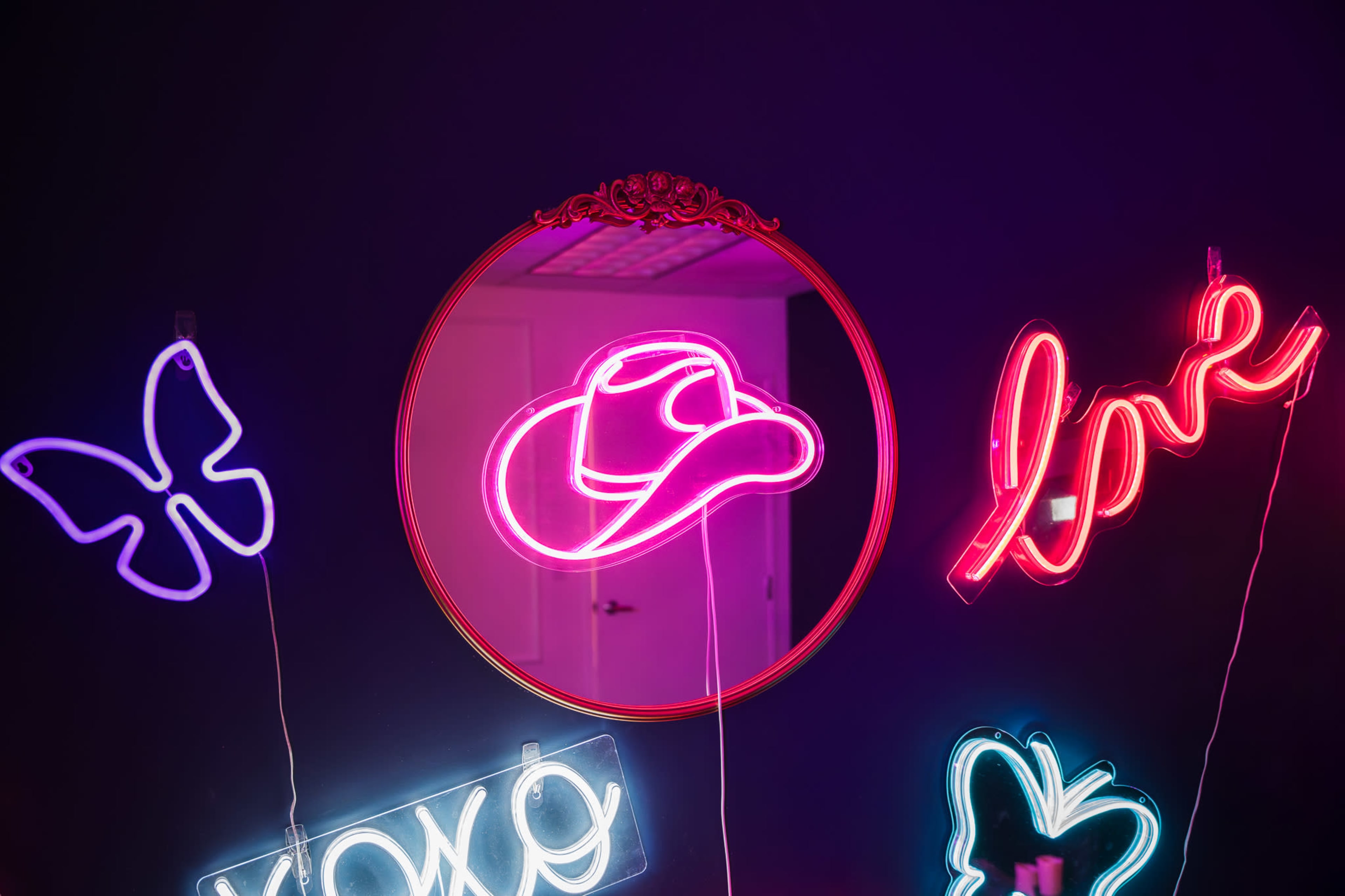 The image features a decorative mirror surrounded by various neon signs, including a cowboy hat, the word "love," a butterfly, and the word "xoxo," all set against a dark background.