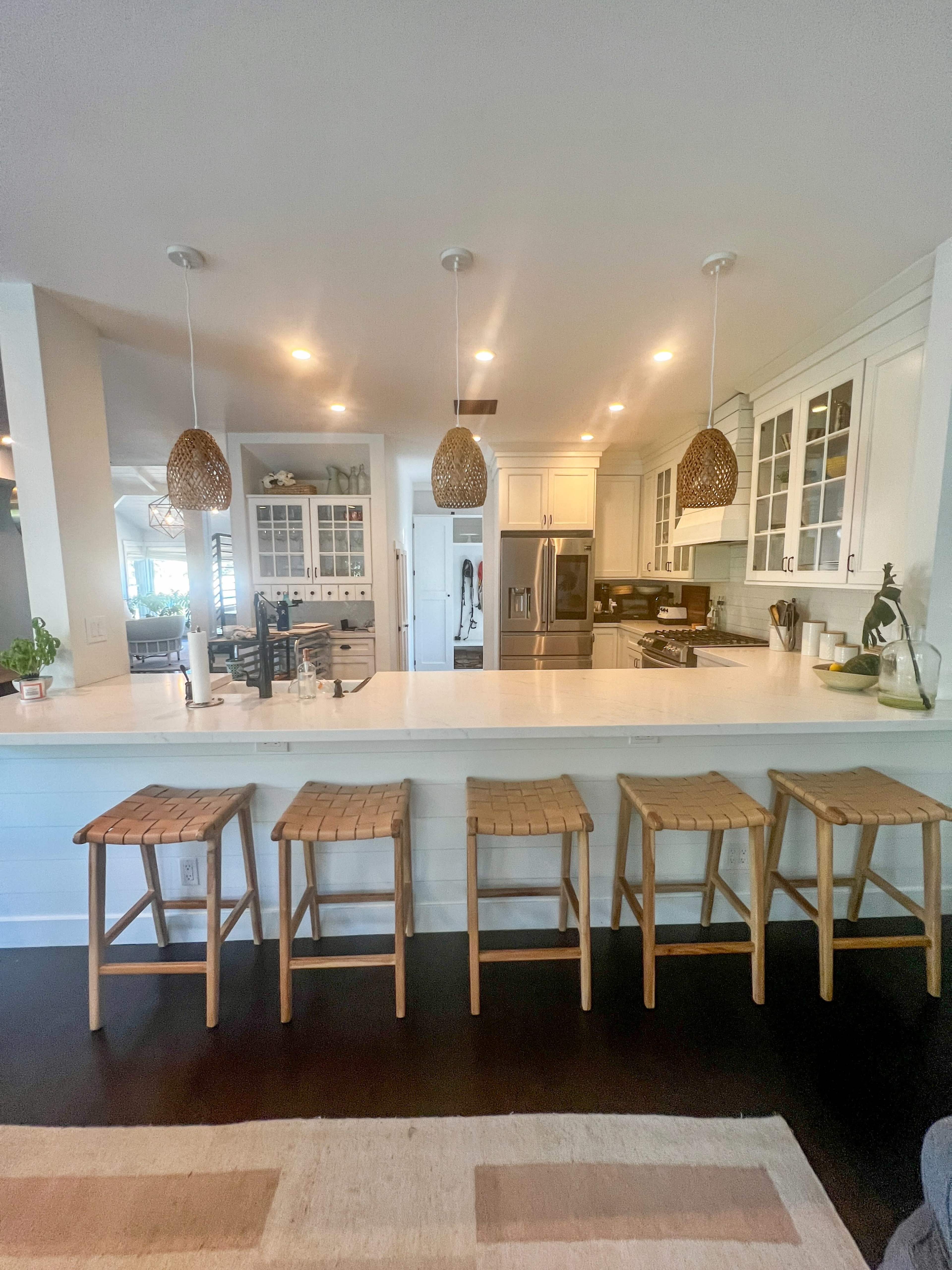 A modern kitchen features a central island with four wooden stools and pendant lighting overhead.